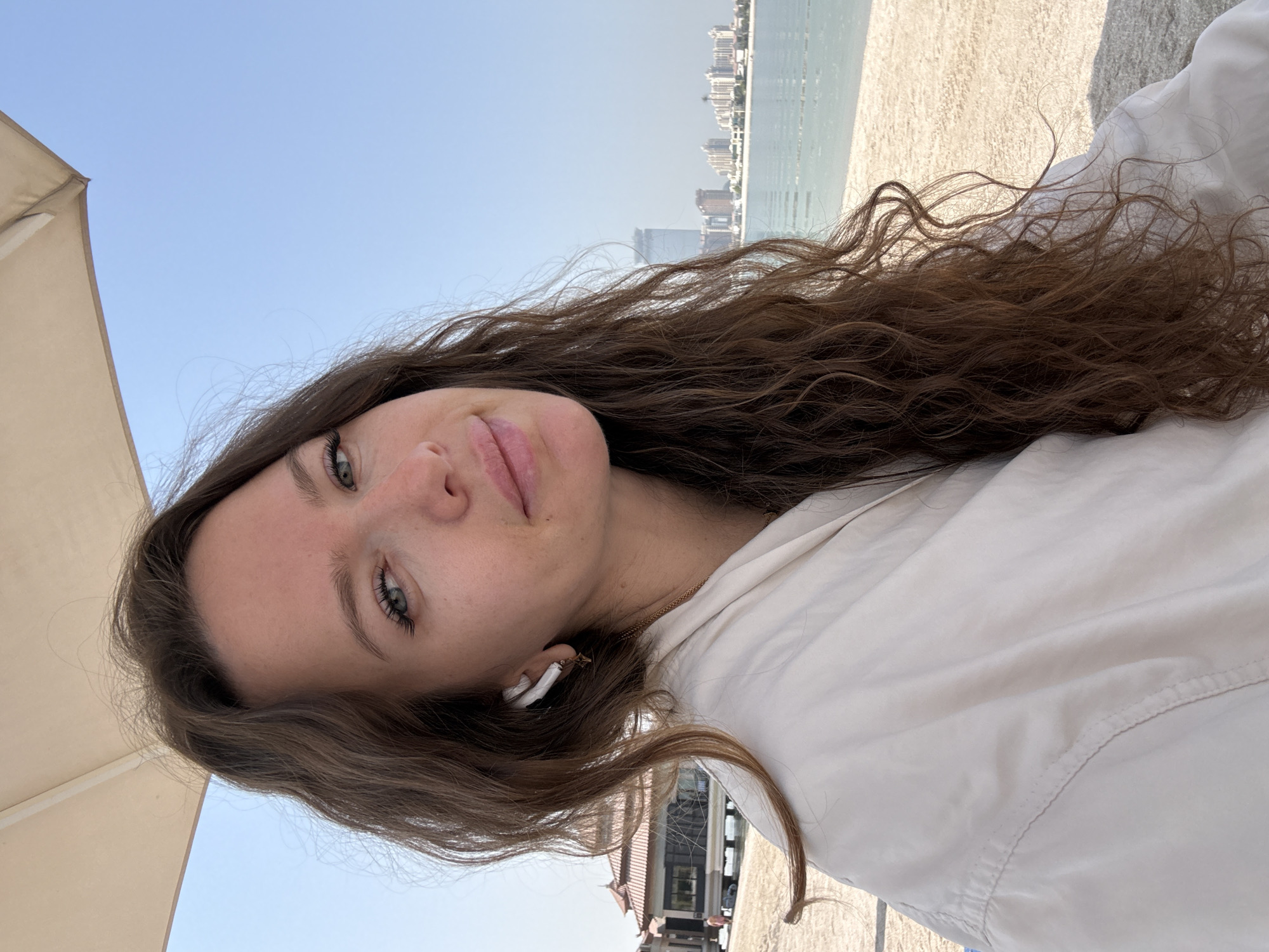 A medium shot photograph captures a woman from the chest up, positioned outdoors. She has long, wavy brown hair, fair skin, and blue-green eyes. The woman is wearing a light-colored jacket and white wireless earbuds. Her gaze is directed slightly upward and to the right, with a subtle smile on her lips.
The background suggests a beach setting in Dubai, United Arab Emirates. To the left, a building with a reddish-brown roof and a darker facade is visible, appearing to be on stilts over water or sand. Further in the distance, along the coastline, a cluster of modern buildings, including high-rises, indicates the urban landscape of Dubai. The sea is calm and light blue, with a few buoys marking an area. The sandy beach stretches from the foreground to the waterline. Above, a portion of a large, light-colored umbrella provides shade. The sky is clear and a bright blue.