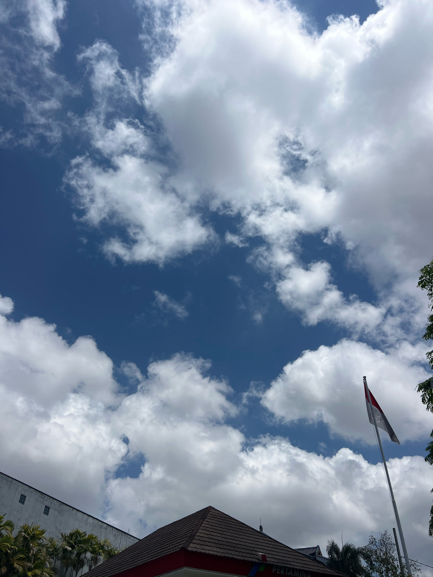 The image shows a clear blue sky with white cumulus clouds. In the foreground, there is a building with a red roof and a white wall with windows. A flag, likely the Indonesian flag given its red and white colors, is flying on a pole to the right of the building. Trees are visible in the lower right and left corners of the image. The building has a sign that says "PERTAMINA", which is an Indonesian state-owned oil and natural gas company, indicating the location is likely a Pertamina gas station or office in Denpasar, Indonesia. The scene appears to be an outdoor setting, under a bright sky, possibly during the daytime.