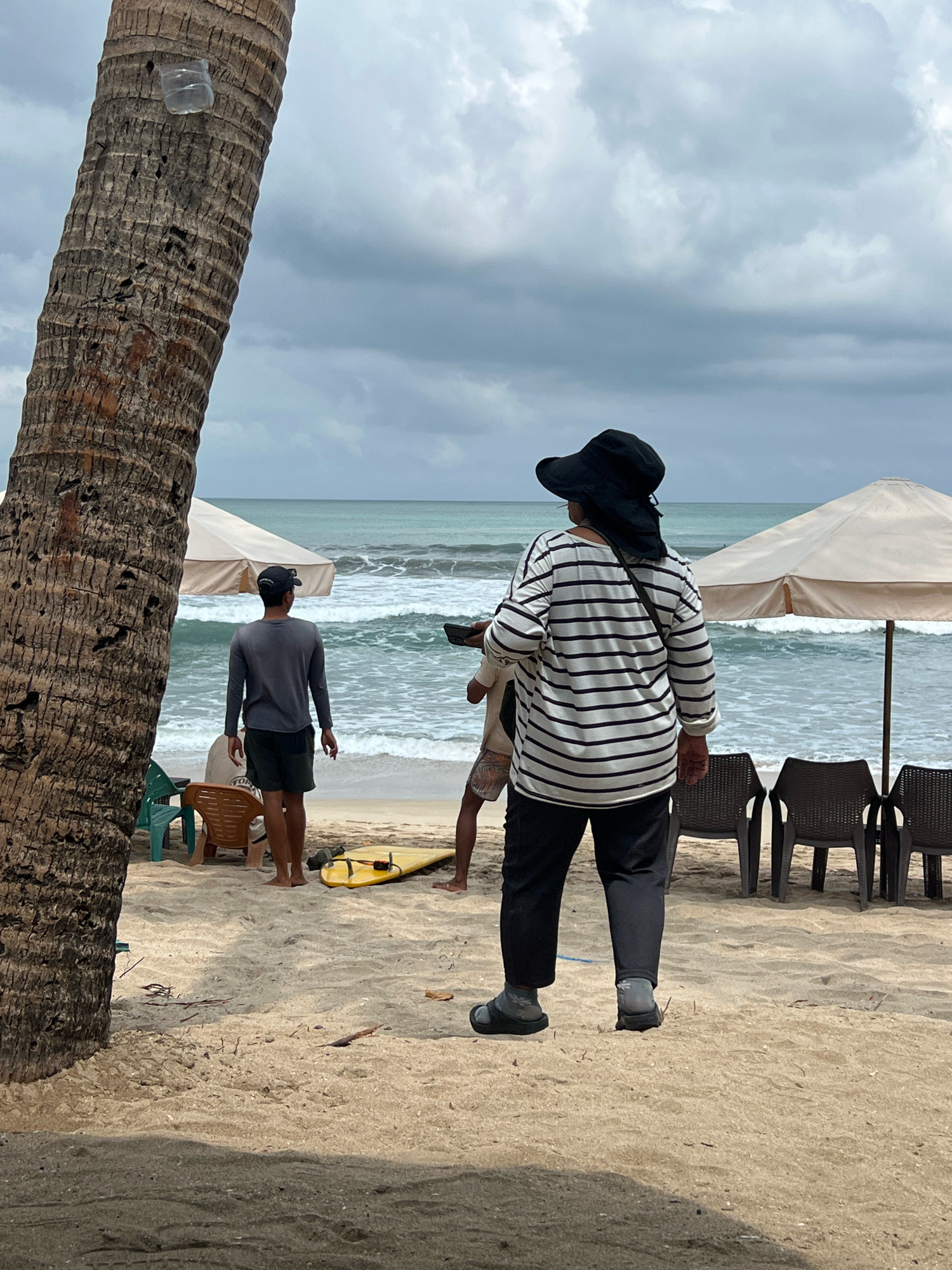The image shows a sandy beach scene in Kuta, Indonesia, on a cloudy day with the ocean and waves in the background. In the foreground, a large palm tree trunk dominates the left side of the frame. A person wearing a black wide-brimmed hat and a horizontally striped long-sleeved shirt stands facing away from the camera, looking towards the ocean. They are wearing dark pants and sandals with socks. To the left of this person, another individual in a gray long-sleeved shirt and dark shorts is also facing the ocean. Between them, a yellow surfboard rests on the sand. In the mid-ground, two beach umbrellas provide shade over empty chairs. The overall atmosphere is casual and relaxed, typical of a beach environment.