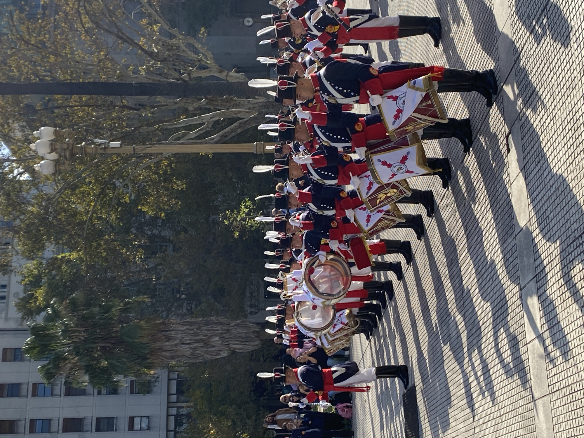 A medium shot of a band, likely a military band, dressed in red, white, and blue uniforms marching on a paved plaza in Buenos Aires, Argentina. The band members are carrying drums. There is a tall lamppost and buildings in the background. Shadows of the band members are cast on the ground, indicating sunny weather. The plaza appears to be near a government building, indicated by the style of architecture.