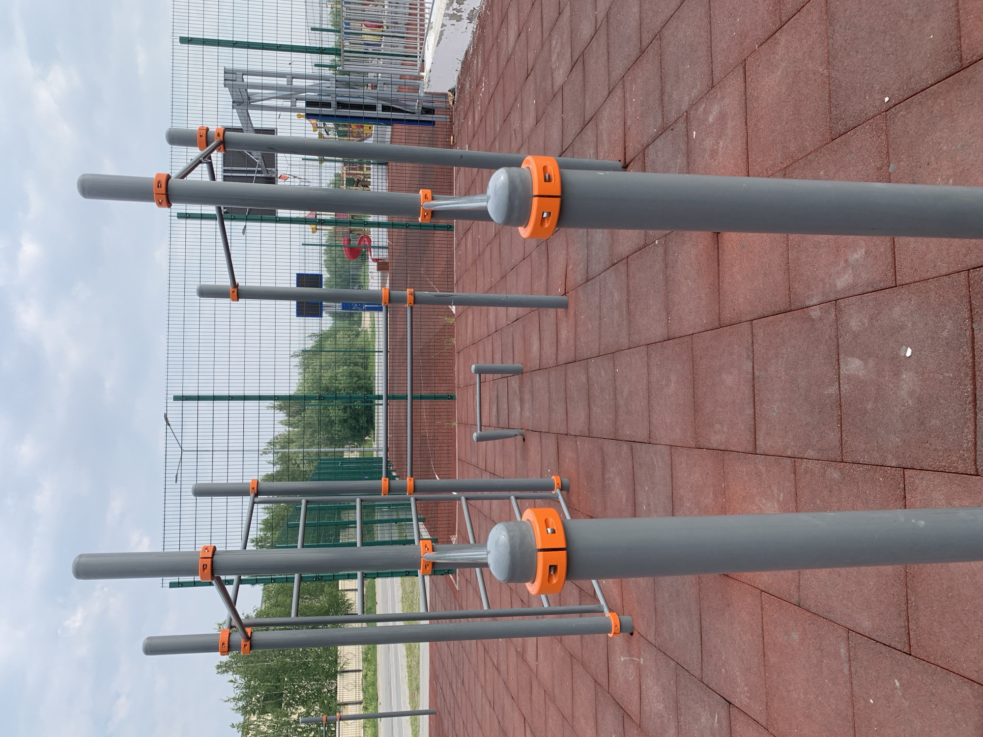This is a medium shot, eye-level, outdoor photograph of an exercise equipment area in the city of Megion, Russia. The ground is covered with red, square rubber tiles. In the foreground, two large, gray cylindrical posts with orange accents rise vertically. The posts are part of a set of outdoor fitness equipment. Behind these, a series of parallel bars, horizontal ladders, and vertical bars form various workout structures. A black metal fence with green framing surrounds the area. Beyond the fence, there are trees and a cloudy sky. In the background, glimpses of a playground with red slides and other equipment are visible. There are no people in the image.