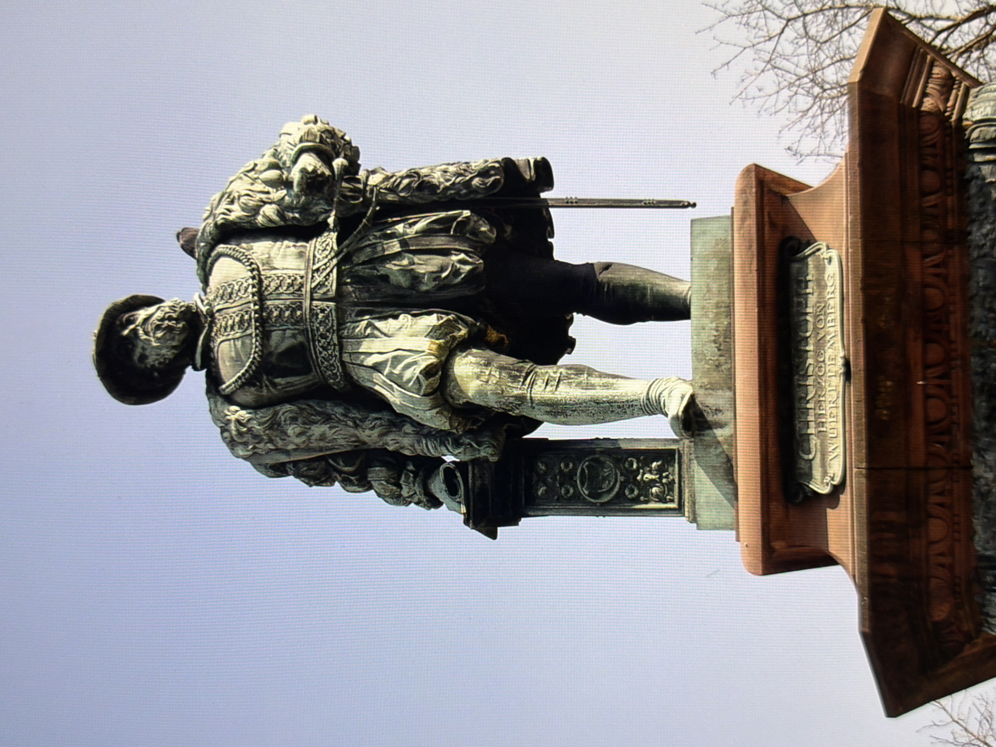 This is a full-body, eye-level shot of a bronze statue of a man wearing historical clothing, a hat, and a beard, with a bird perched on his shoulder. The statue is of Christoph, Duke of Württemberg, and it is located in the city of Kyiv, Ukraine. The statue stands on a large, multi-tiered pedestal made of stone. The background is a clear, pale blue sky. The lighting suggests it is daytime, likely sunny. The statue is made of aged bronze, with a green patina visible. The man in the statue is depicted in a formal pose, looking slightly upwards, with one leg forward. He is holding a sword in his right hand, which hangs down his side. The pedestal has inscriptions on it. The overall impression is of a historical monument.