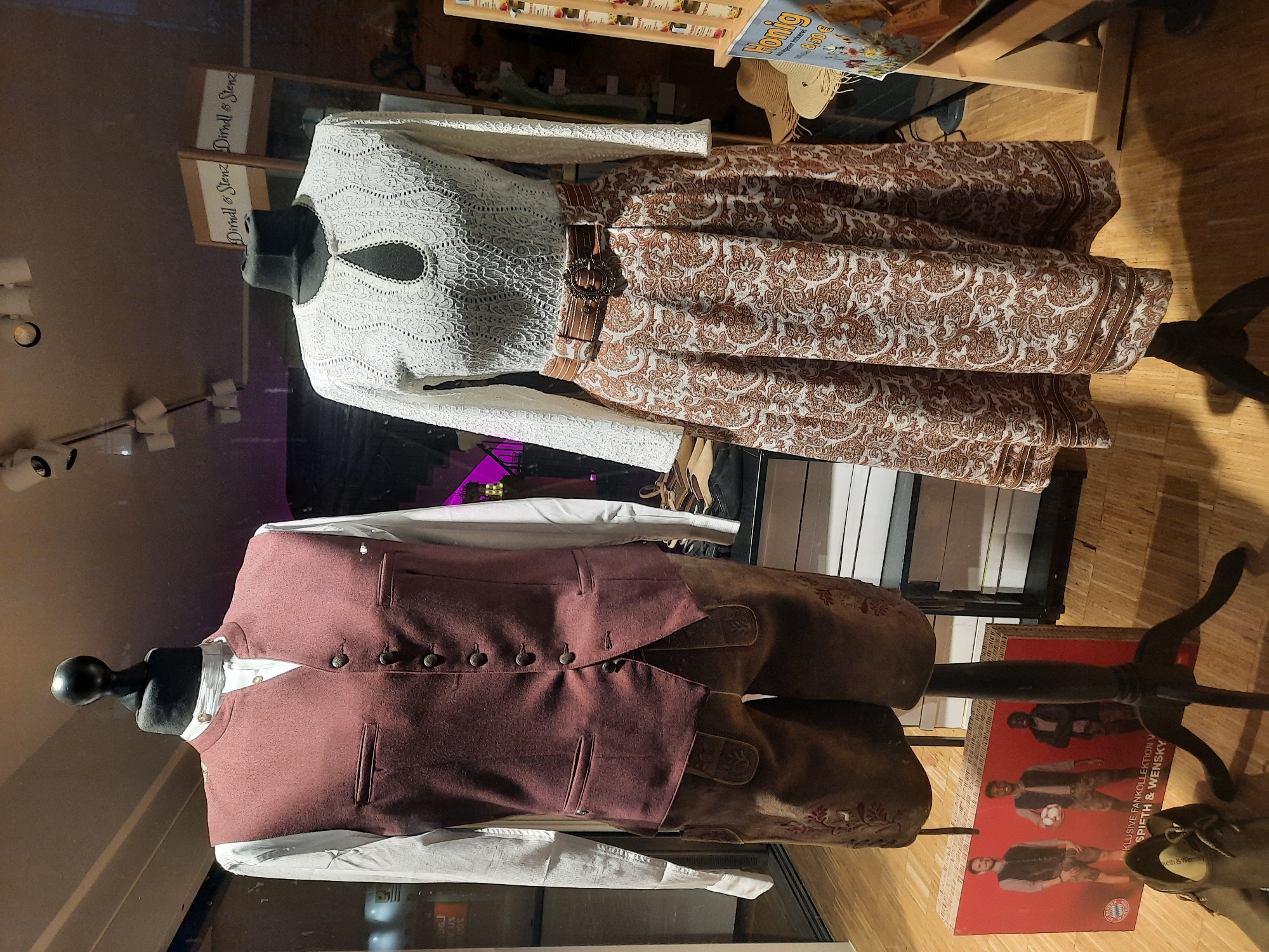 The image shows a shop window display featuring traditional Bavarian clothing. On the left, a male mannequin wears a white collared shirt, a maroon vest with buttons, and brown leather shorts with intricate embroidery. On the right, a female mannequin is dressed in a white lace top with a keyhole neckline and a long, brown and white patterned skirt cinched with a brown leather belt. Behind the mannequins, several wooden signs with "Dirndl & Stenz" are visible. A red cardboard display featuring three men in similar traditional attire is placed on the floor in front of the male mannequin. The shop appears to be located in Rosenheim, Germany, as suggested by the presence of Bavarian clothing and potentially local business signage. The lighting suggests an indoor setting, possibly a boutique or clothing store.