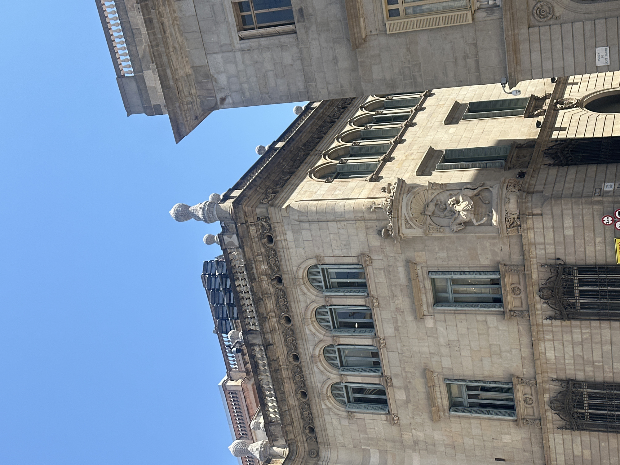 The image shows a street view in Barcelona, Spain, likely in the Gothic Quarter or El Born district, given the architectural style. The primary subject is the exterior of two ornate, classical-style buildings bathed in bright sunlight. The building in the foreground is a light-colored stone structure with multiple floors, characterized by arched windows with green shutters, decorative cornices, and intricate stonework. A notable feature on the corner of this building is a sculpted relief of a knight on horseback. Above the main facade, the building's roofline is adorned with decorative elements and what appear to be air conditioning units or similar rooftop equipment. To the right, a portion of another stone building is visible, also featuring classical architectural details and windows with shutters. The sky above is a clear, deep blue. Street signs are visible at the bottom left, including a sign indicating a pedestrian zone and another with a red circle and diagonal bar over a "2.5m" symbol, suggesting a vehicle height restriction. A street name sign reading "PLAÇA DE SANT JAUME" is visible on the right building. There are no people clearly visible in the image. The overall impression is one of historical architecture in a well-maintained urban setting.