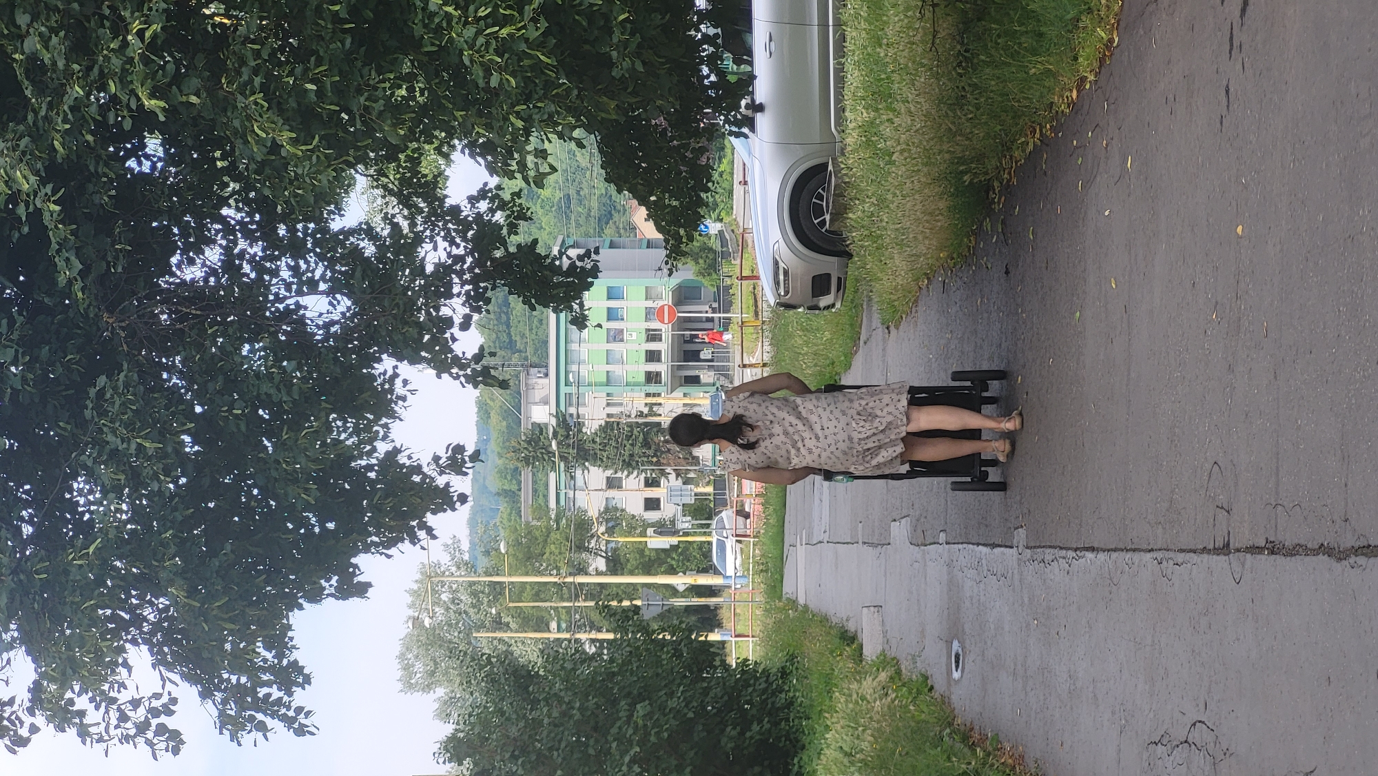 A woman with her back to the camera is walking on a paved path, pushing a stroller. She is wearing a floral print dress and sandals. The path is bordered by grass and foliage. In the background, there is a silver SUV, a light green building with multiple windows, trees, and utility poles. The scene appears to be in an urban or suburban area of Prešov, Slovakia.