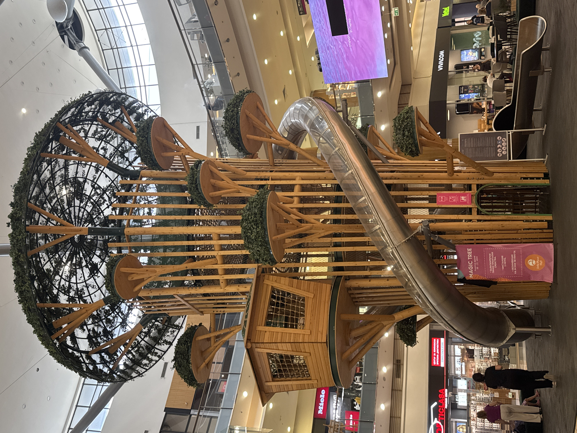The image depicts a large, tree-shaped play structure in a shopping mall. The structure is made of wood and metal, with a spiral slide and various platforms and a small enclosed wooden cabin. The play structure is decorated with artificial greenery. In the foreground, a pink sign with text in Bulgarian is visible, indicating it's part of Delta Planet Mall. Two women are walking in the lower left portion of the image, and other people are seated at tables in the background. The mall environment includes shops with illuminated signs, including "TIGLASA" and "VIVACOM." The overall setting suggests an indoor public space, likely a shopping mall in Varna, Bulgaria, given the mall name.