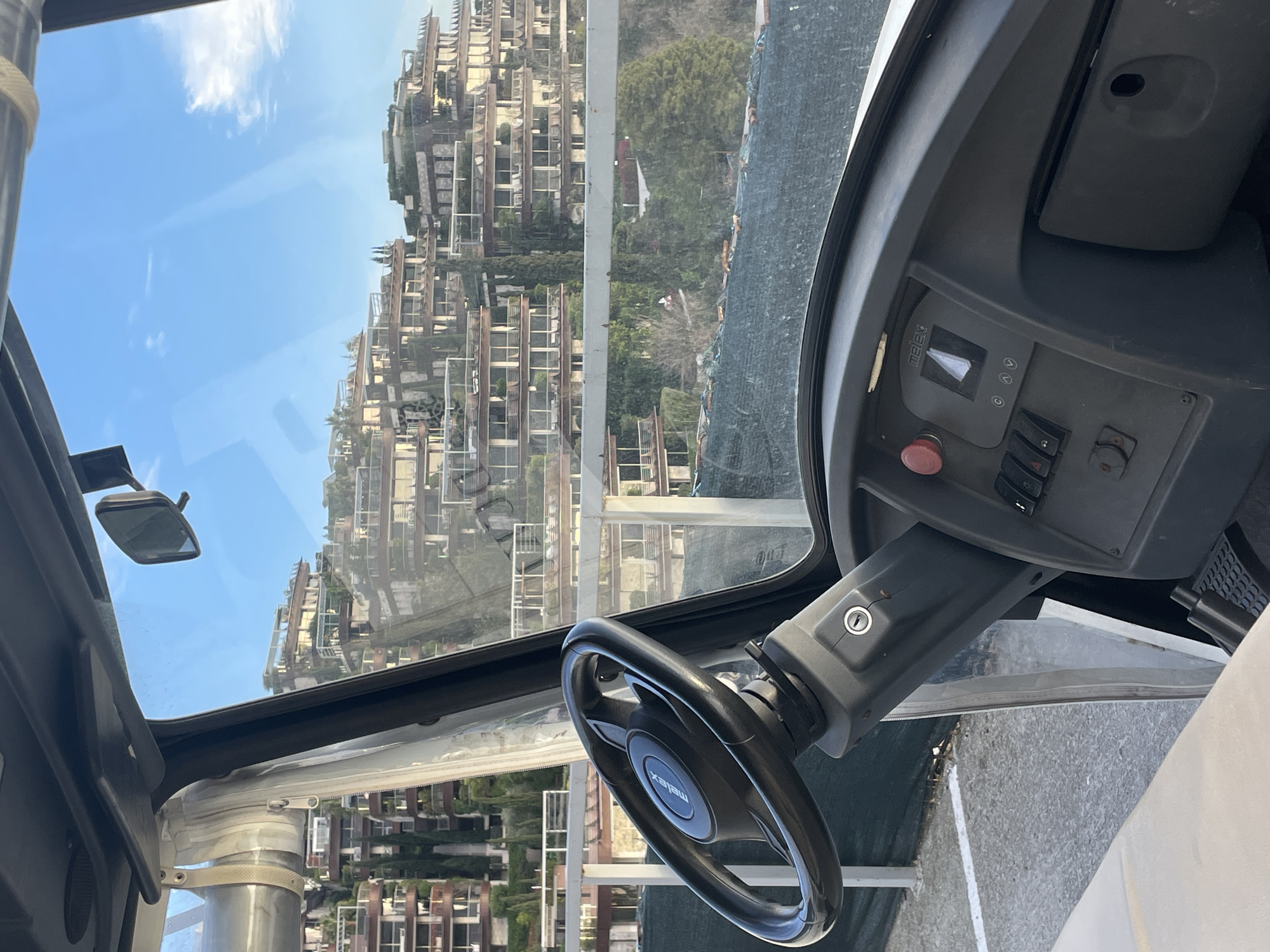The image displays an exterior scene viewed from inside a stationary electric utility vehicle, identified by the "melex" branding on its black steering wheel and dashboard. The vehicle's interior components visible include the steering wheel positioned left of center, a dashboard with an ignition keyhole, a red emergency stop button, a digital display indicating "melex" and directional arrows, and multiple black toggle switches. A portion of a beige seat is visible on the lower left, and a black rubber pedal appears on the lower right floor. A rearview mirror is affixed to the upper right section of the windshield frame.
Outside the vehicle, a white metal railing spans horizontally across the mid-distance. Below this railing, a dark green mesh netting covers a sloped area containing scattered trees and vegetation. The immediate foreground outside the vehicle reveals a paved surface with faint white ground markings.
The background features a steep hillside densely covered with large, multi-story, terraced buildings. These structures possess numerous balconies and flat roofs integrated with green landscaping and plantings. Tall, slender evergreen trees are interspersed among the buildings and on the surrounding slopes. The sky above is clear blue with sparse white clouds.
This location is identified as the **Dukley Gardens** residential complex on the Zavala Peninsula in Budva, Montenegro, based on the distinctive terraced architecture and hillside setting. No individuals are depicted, and no specific actions or interactions are occurring.