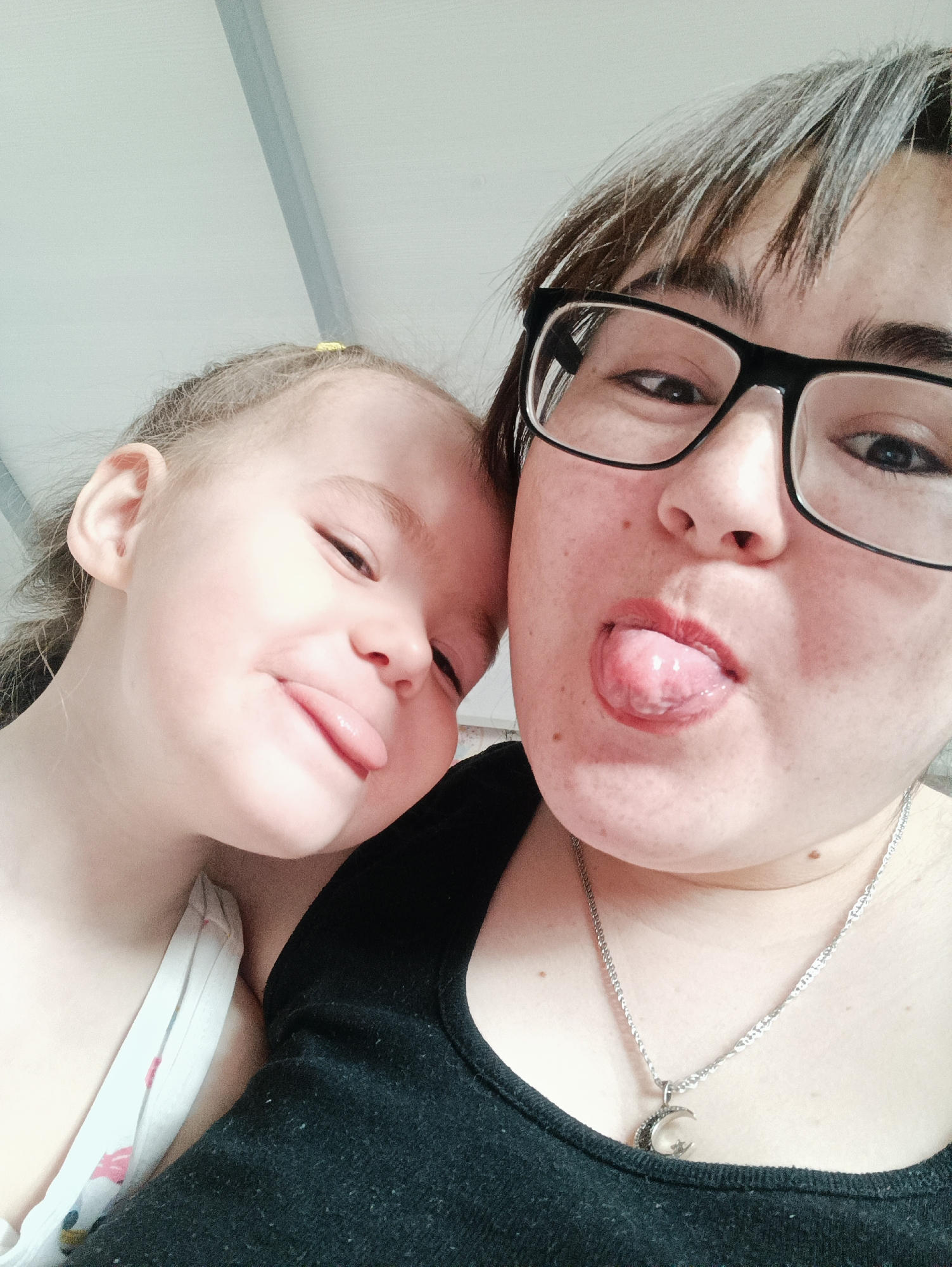 This close-up portrait features an adult woman and a young child, likely a mother and daughter, captured in a playful moment in Noviy, Russia. The adult is positioned on the right side of the frame, wearing black-framed glasses and a black tank top. Her dark hair, which shows some visible grey strands, is styled with bangs. She has a silver necklace with a crescent moon-shaped pendant. The child is on the left, with fair skin and lighter hair, secured with a small yellow hair tie. She is wearing a white top with a faint pattern.
Both individuals are making funny faces, sticking out their tongues towards the viewer, conveying a sense of joy and lightheartedness. The adult's tongue is visibly extended, and her cheeks show a scattering of freckles or moles. The child's tongue is also out, and her eyes are squinted in amusement.
The background is a simple, light-colored wall or ceiling, with a thin grey strip visible in the upper part of the image, suggesting an indoor setting. The lighting is bright and even, indicating daytime or well-lit interior conditions. There is no visible text in the image. The overall mood is affectionate and fun.