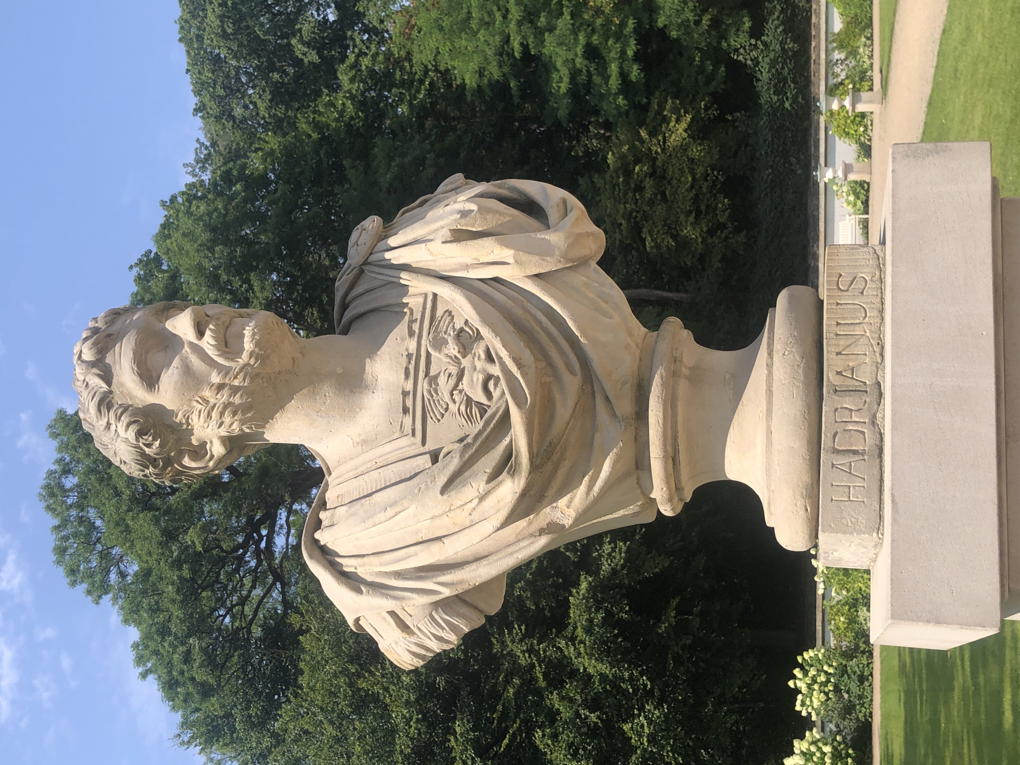 The image displays a close-up view of a stone bust of a man, identified by the inscription "HADRIANUS" on its base. The bust is positioned outdoors in a park-like setting, with lush green trees forming the background. The sky above is a clear blue with a few scattered clouds. The bust itself is detailed, showing a bearded face with curly hair and wearing draped clothing, possibly a toga or a military uniform. The stone appears weathered, suggesting it is an old monument. The inscription is carved into a rectangular stone block at the base of the bust. In the lower portion of the image, a well-maintained lawn and a pathway are visible, with hints of decorative bushes and possibly other statues or urns in the distance. The overall scene suggests a historical or memorial site. This bust is located in the Łazienki Park in Warsaw, Poland, specifically in front of the Palace on the Isle.