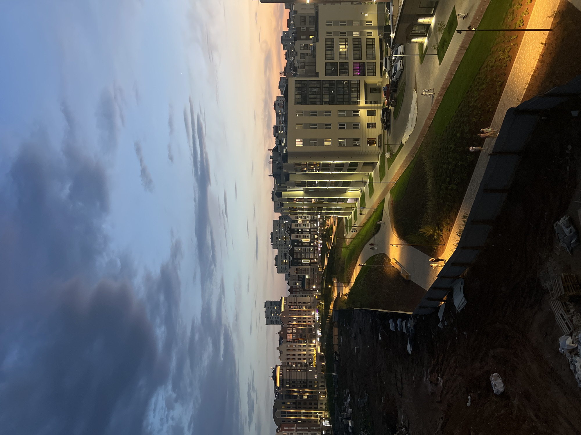 Captured from an elevated vantage point, the image presents an urban landscape at twilight in Pervomaysky, Russia. Modern, multi-story residential buildings, illuminated with interior and streetlights, stretch into the distance under a sky transitioning from blue to orange hues. A green strip of manicured grass and walkways divides the buildings. A construction site is visible in the foreground, indicating ongoing development. People can be seen walking along the paths and sidewalks. Cars are parked beside the buildings. The lighting indicates the scene is taking place at night.