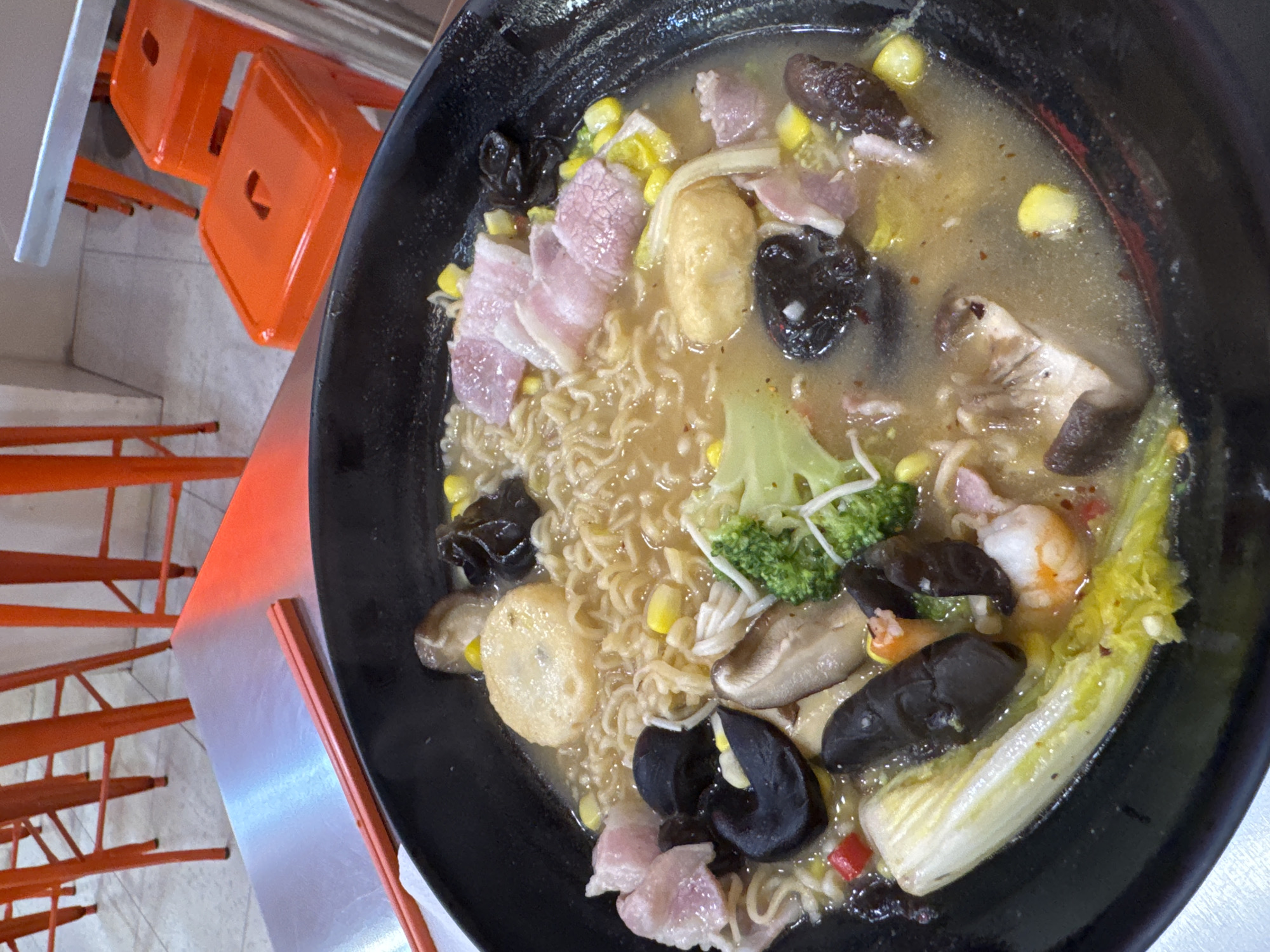 A bowl of ramen sits on a metal surface next to a pair of red chopsticks. The ramen contains noodles, broth, corn, meat, broccoli, leafy greens, and mushrooms. Nearby are stacked orange chairs, and the scene appears to be indoors. No people are directly visible. The image suggests a dining setting. The city and country are provided as background information, but do not directly contribute to the visual contents of the image.