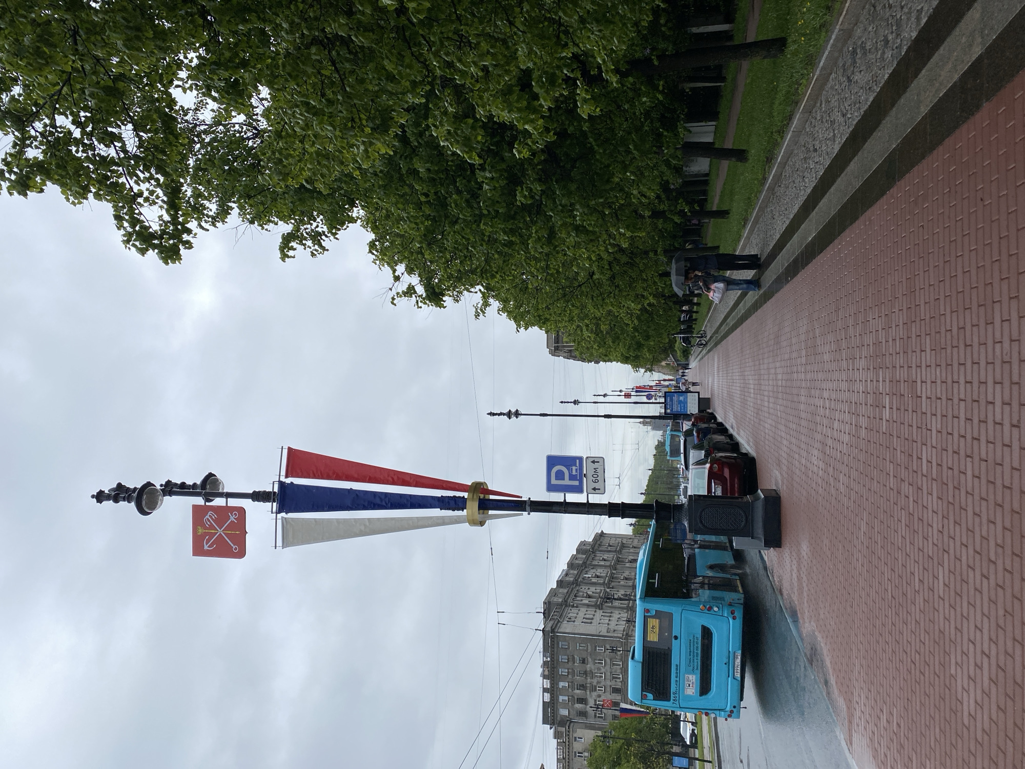 The image depicts a street scene in Saint Petersburg, Russia, likely on a rainy day. A long, red brick sidewalk runs alongside a street filled with parked cars and a blue bus. Trees line the sidewalk. People are visible under an umbrella further down the sidewalk. A lamppost decorated with the colors of the Russian flag (red, white, and blue) stands prominently in the foreground. A parking sign is also visible. Buildings line the opposite side of the street. The sky is overcast.