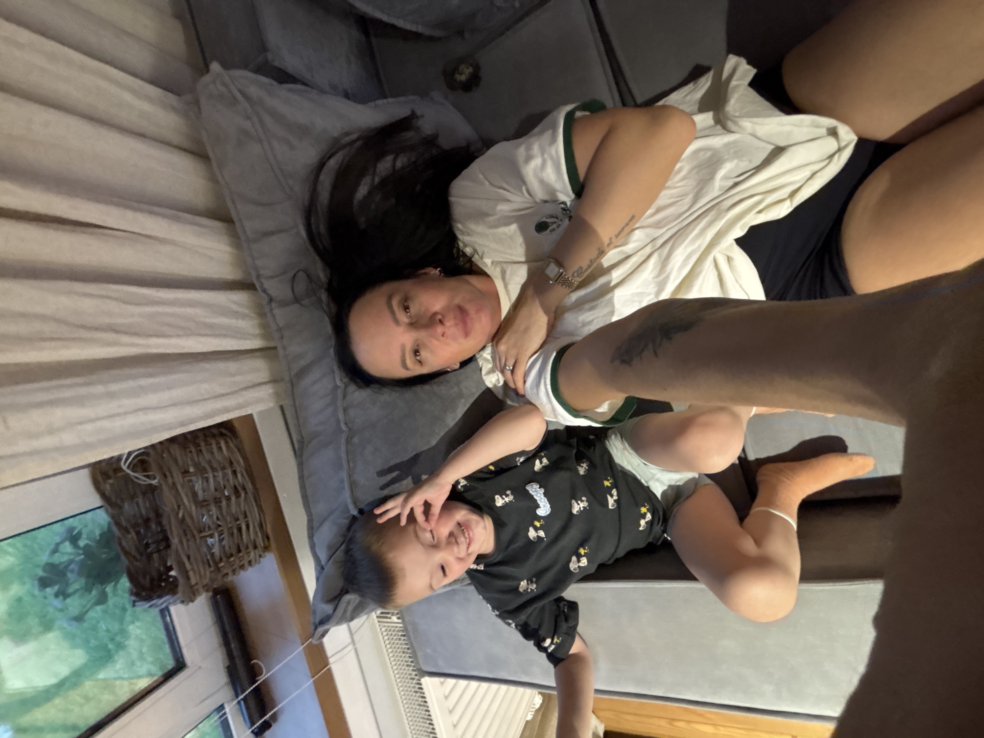A woman and a child are lying on a gray sofa indoors. The woman has long dark hair and is wearing a white t-shirt with green trim and dark bottoms. The child, appearing to be a toddler, is wearing a black t-shirt with cartoon images. They are both facing upwards towards the camera. A window with a view of greenery is visible on the left. A woven basket hangs near the window. Blinds cover a nearby window. The location is unspecified, and no evidence suggests it is in Chaikovsky, Russia.
