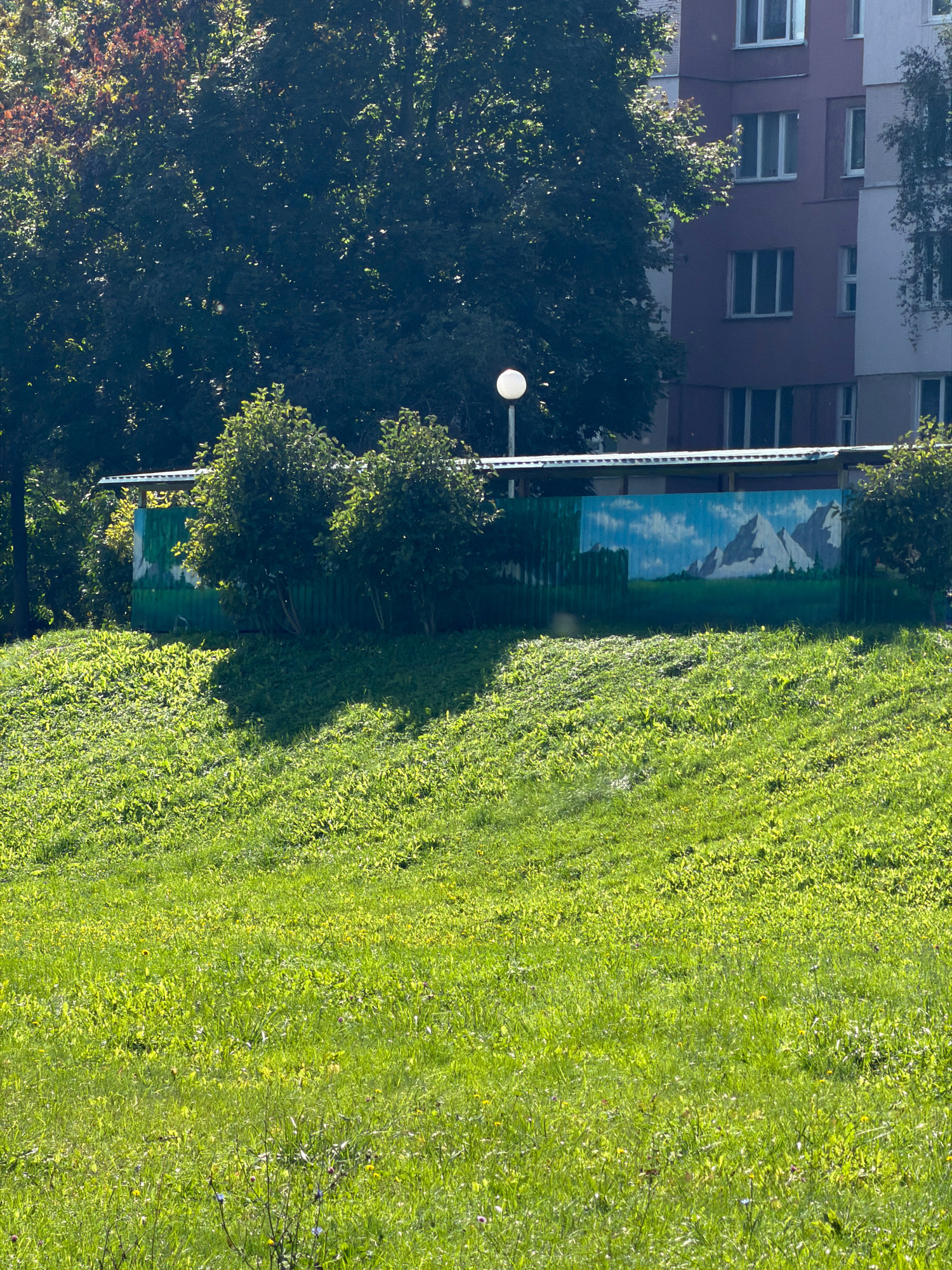 The image shows a grassy hill with a painted fence in the background. The fence is painted with a mountain landscape. Behind the fence is a building with multiple windows. The scene appears to be in an urban environment, possibly a park or a residential area. The grass is green and lush, suggesting it is springtime or summer. There are no people or animals visible in the image. The lighting suggests it is daytime, with the sun casting shadows on the grass. The overall atmosphere is peaceful and serene.