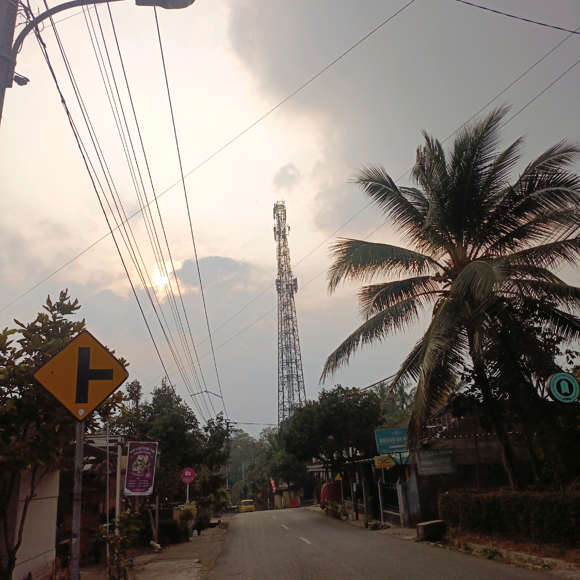 The image depicts a street scene in Salatiga, Indonesia, with a cellular tower prominently visible in the background. The sky is overcast with the sun partially obscured by clouds, creating a diffused light. In the foreground, a yellow traffic sign with a black symbol indicating a T-junction is mounted on a pole. To the right of the sign, a lush palm tree with visible coconuts partially obstructs the view of the sky. Further down the street, several buildings and shops line the road, one of which has a sign that reads "Warung Band Pay". A yellow car is parked on the side of the road. Utility wires crisscross the scene, strung between poles and obscuring parts of the sky and tower. The overall impression is of a suburban or semi-urban environment.
