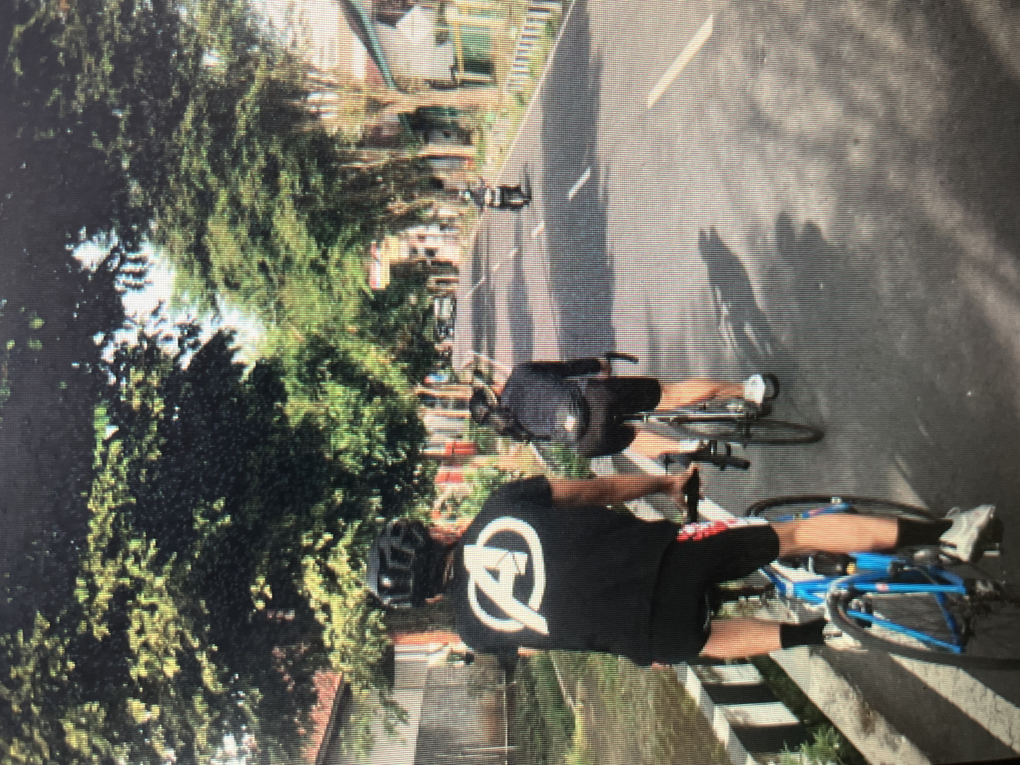 The image depicts a sunny street scene in Sejahtera, Indonesia, with two individuals cycling and another on a scooter. In the foreground, a person wearing a black t-shirt with a white Avengers logo and a black helmet is riding a blue bicycle. They are wearing black cycling shorts and dark socks. To their right and slightly ahead, another individual on a bicycle, seen from behind, is wearing a black jacket and black shorts. Further down the road, a scooter is being ridden by a person dressed in dark clothing. The street is lined with trees casting shadows, and buildings are visible on either side. The road has white lane markings.