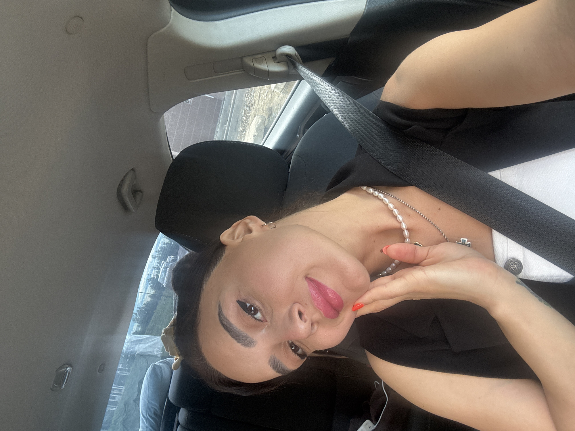 The image shows a woman taking a selfie inside a car, likely in Tbilisi, Georgia. She has dark hair pulled back and is wearing a black vest over a white top. She is also wearing a pearl necklace with a cross pendant and has bright red nail polish. The car interior is visible, including a seatbelt and the headrests of the seats. The background outside the car shows a mix of buildings and greenery, suggesting an urban or suburban environment. The lighting appears to be natural daylight.
