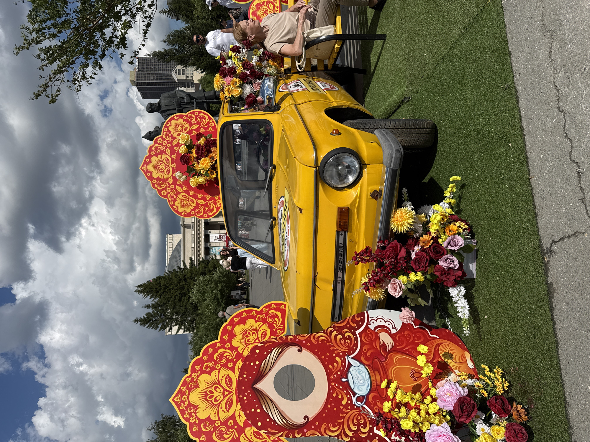 The image captures an outdoor event in Novosibirsk, Russia, likely a city celebration or exhibition, given the festive decorations. A bright yellow vintage car, possibly a ZAZ model, is prominently displayed on artificial grass. The car is adorned with large, colorful decorative cutouts resembling Russian folk art and abundant floral arrangements, primarily in shades of red, yellow, and pink. These flowers are clustered around the car and on its roof, suggesting a summer event.
To the left of the car, a large, flat cutout of a matryoshka doll, with a face cut out for photo opportunities, is positioned, also decorated with similar folk art patterns. To the right of the car, a woman is seated on a yellow bench, looking away from the camera. She is wearing a beige top and pants and has a light-colored bag next to her. In the background, other people can be seen mingling, and a statue of two figures is visible behind the car, partially obscured by floral arrangements. A tall, modern building with a sign that appears to say "СИТИЦЕНТР" (City Center) stands in the distance, indicating an urban setting. The sky is partly cloudy with dramatic, white cumulus clouds against a blue backdrop. The overall scene suggests a public gathering with a cultural or artistic theme.