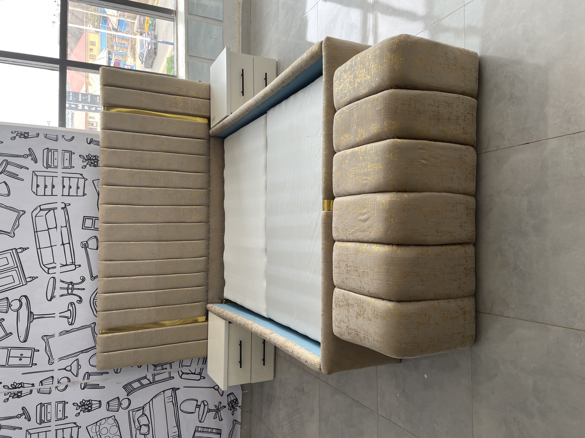 This image captures a modern bedroom or furniture showroom setting, showcasing a stylish bed and its immediate surroundings. The scene is static, devoid of people.
The central subject is a large bed featuring an upholstered frame, headboard, and footboard. The upholstery is a light beige or champagne color, subtly textured with a gold, distressed pattern. The headboard consists of several horizontal, flat padded segments, accented by a thin, polished gold metallic strip running across its width. The footboard mirrors this design with horizontal padding, but its segments appear thicker and more rounded. A white mattress rests within the frame, and a thin strip of light blue fabric is visible underneath the mattress, along the frame's interior edge.
Flanking the bed are two small, white, glossy nightstands or storage units, each with two black handles, appearing to be integrated with or positioned tightly against the bed frame.
The wall behind the headboard is adorned with distinctive white wallpaper featuring numerous black line drawings of various furniture pieces, including chairs, tables, dressers, lamps, and potted plants, creating a playful and artistic backdrop.
To the left of the bed, a large window offers a view of an outdoor urban scene during daylight hours. Visible elements outside include buildings with red roofs, power lines, and what appear to be vehicles, including a yellow auto-rickshaw-like vehicle and a grey car. Faint text on an outdoor building through the window seems to read "SALON" and "GYM." The floor and the wall to the right of the bed are covered with light grey square tiles.
The overall impression is one of modern design and comfortable aesthetics, likely in Gura Top, Nigeria, given the contextual information.