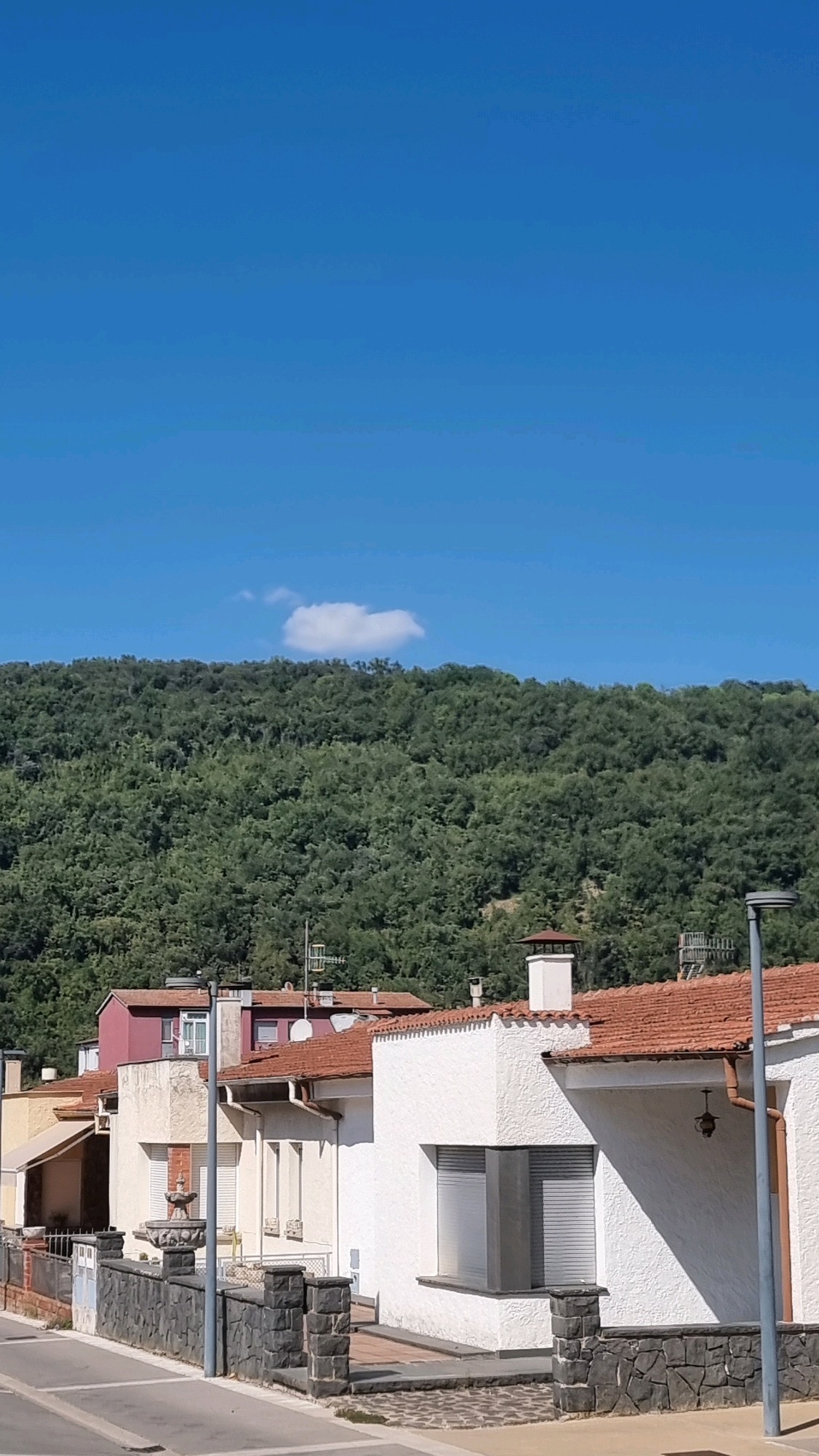 The image depicts a view of houses and a forested hill under a clear blue sky with a single small cloud. The foreground features a street or road, and a low stone wall with pillars separates the properties from the street. Several houses with terracotta roofs are visible, some painted white, one with a red roof and pinkish walls, and another a pale yellow. One white house has prominent gray shutters on its windows. A street lamp is present on the right side of the image. The background is dominated by a densely wooded hillside. The scene appears to be a residential area within or near Sant Joan Les Fonts, Spain.