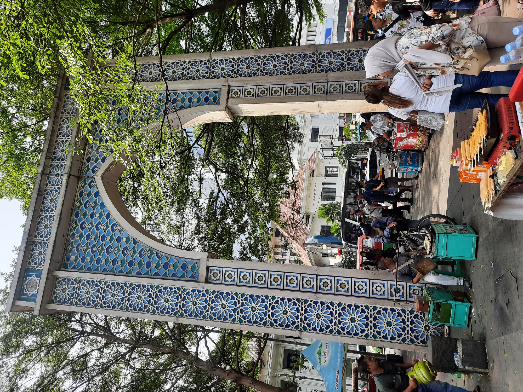 The image depicts an outdoor scene featuring a large, ornate archway decorated with blue and white geometric patterns. In front of the archway, several people are gathered. To the left, a man in a dark green shirt carries a bright green gas cylinder. In the center, a man sits on a stool next to a bicycle and two turquoise food stalls. He appears to be preparing or selling food. To the right, two women are standing and talking, one wearing a white headscarf and the other a white t-shirt. Another man is leaning on the bicycle. Behind them, various food carts and a bus are visible. The environment suggests a bustling street or market area. Based on the presence of a "Le Minerale" branded tent and Indonesian text on the food carts, the location is likely in Indonesia. Further context identifies the location as the Asian-African Conference Museum (Museum Konferensi Asia Afrika) in Bandung City, Indonesia, which is known for this distinctive archway.