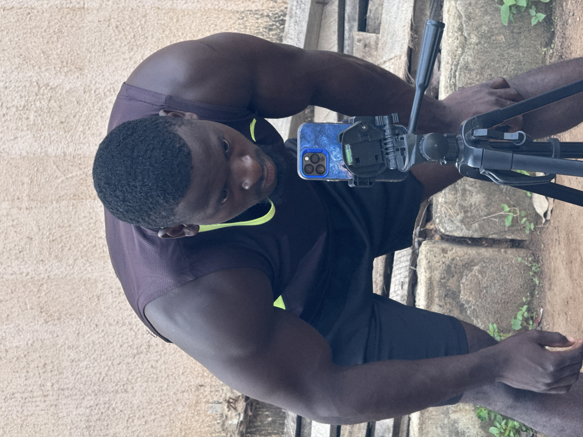 The media file shows a young man, possibly in his late teens or early twenties, taking a selfie with a tripod and camera. He is wearing a dark purple athletic tank top with a neon yellow trim and dark shorts. The background suggests an outdoor setting, with rough, textured surfaces that could be concrete or stone. There are also some hints of greenery, indicating the presence of plants. The lighting appears to be natural daylight, suggesting the photo was taken during the day.
The man is looking directly at the camera, and his expression is neutral, perhaps slightly focused on the act of taking the photo. The tripod is positioned in front of him, with the camera mounted on it, angled to capture his reflection. A smartphone with a decorative case, featuring a swirling blue pattern, is visible attached to the tripod. The man's muscular physique is apparent, especially his arms and shoulders.
The scene appears to be in a somewhat rustic or undeveloped environment, based on the rough textures and the presence of natural elements. There is no visible text in the image. The overall impression is that of a casual, personal moment being captured.