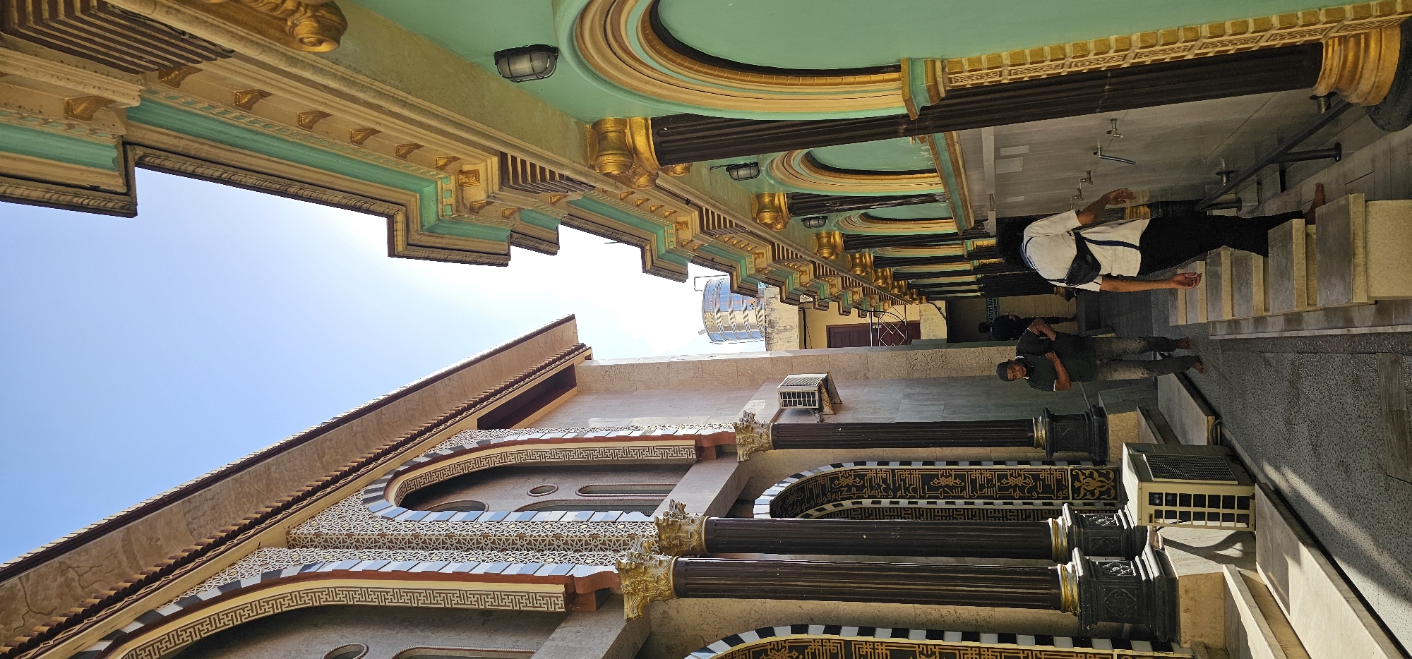 This photo depicts a building in Denpasar, Indonesia. The building has ornate architecture with gold, turquoise, and beige detailing, decorative geometric patterns, arched windows, and a tiled roof. In the foreground, there is a colonnade with dark pillars and gold accents. Two barefoot men are visible. One man is wearing a white shirt with a black bag. The other man is wearing a dark green shirt. They appear to be on a stairway, and the man in green is walking downward. The sky above is clear and blue.