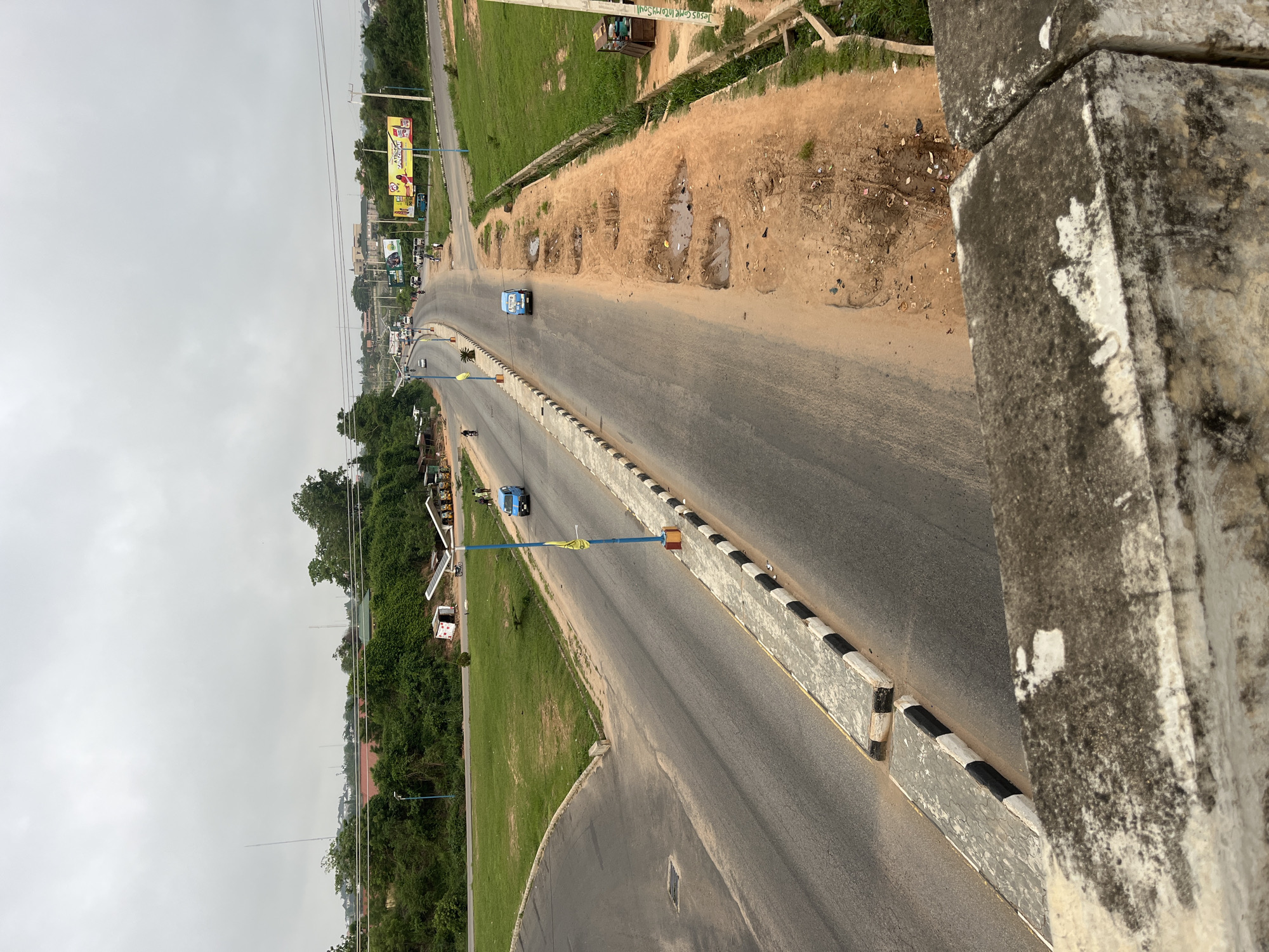 A ground-level, long shot shows a two-lane road in Osogbo, Nigeria, under an overcast sky. The road stretches toward the horizon, with vehicles visible in the distance. Roadside barriers marked with alternating black and white stripes run along the road's edge. To the left of the road, there is a grassy area with trees and buildings. To the right of the road, the terrain is sandy and uneven, with some exposed soil. In the background, billboards and other structures are visible, with power lines overhead. The foreground features a concrete structure from where the shot was taken.
