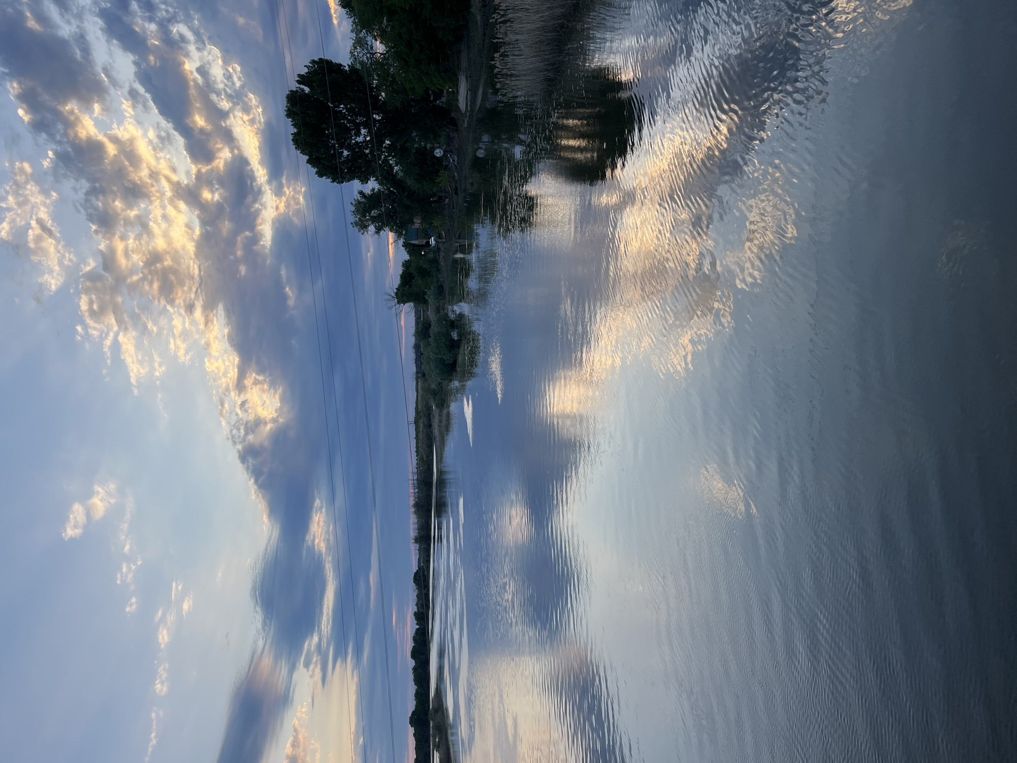 This landscape photograph captures a serene waterscape at dusk in Krasnyye Barrikady, Russia. The scene is dominated by a large body of water, likely a river or lake, reflecting the dramatic evening sky. The sky is filled with a mix of deep blues and oranges, indicating the sun has recently set or is about to rise. Wispy clouds, illuminated by the lingering sunlight, create a beautiful contrast against the darkening sky and its reflection on the water's surface.
The left side of the image shows the sky with its colorful clouds and a few dark, thin power lines running diagonally. On the right and bottom portions of the image, the rippling water takes center stage, with its surface mirroring the sky's hues and textures. The gentle waves distort the reflections, creating an abstract and painterly effect.
Along the distant bank, a dense line of trees and vegetation is visible, casting dark silhouettes against the brighter sky. The trees appear lush and green, suggesting a temperate season. A subtle hint of human presence can be discerned in the form of some structures and what might be a vehicle near the tree line, peeking through the foliage.
The overall mood of the photograph is tranquil and contemplative, evoking the peacefulness of nature at twilight. The interplay of light and shadow on the water and in the sky creates a visually captivating and atmospheric image, characteristic of a calm evening in a rural or natural setting. There is no visible text in the image.