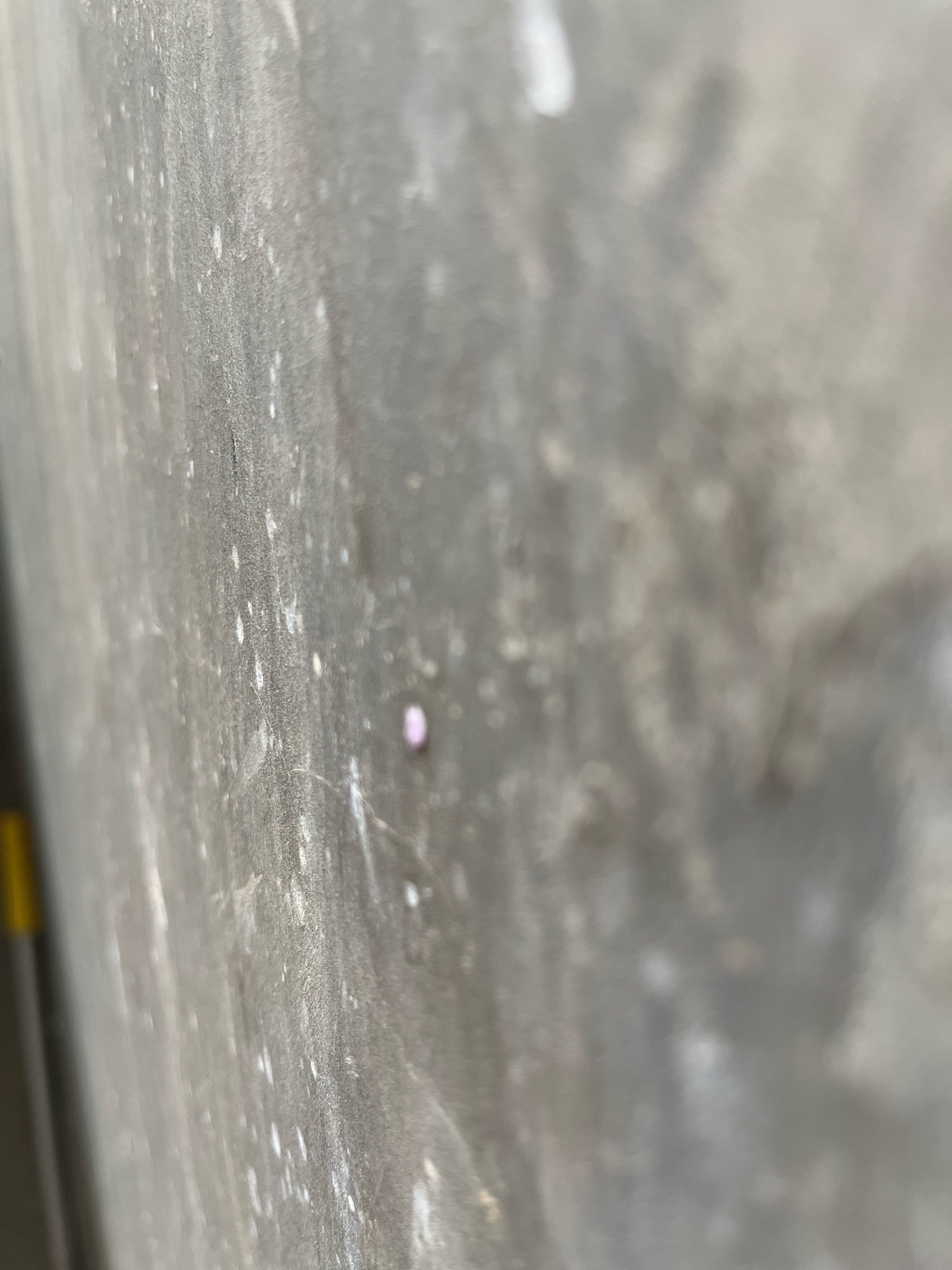 The image shows a close-up, angled view of a textured, grayish-brown surface, possibly concrete or a similar material. The surface appears weathered and dirty, with numerous small white specks and some faint vertical lines. In the center of the image, there is a very small, isolated, pinkish-purple speck. The lighting is uneven, creating subtle variations in shadow and highlight across the surface. The background is out of focus, but a blurred yellow object and a dark vertical line are visible on the left side of the frame. There are no people, actions, or interactions depicted in the image. The location in Kebumen, Indonesia, is not visually identifiable from the provided image.