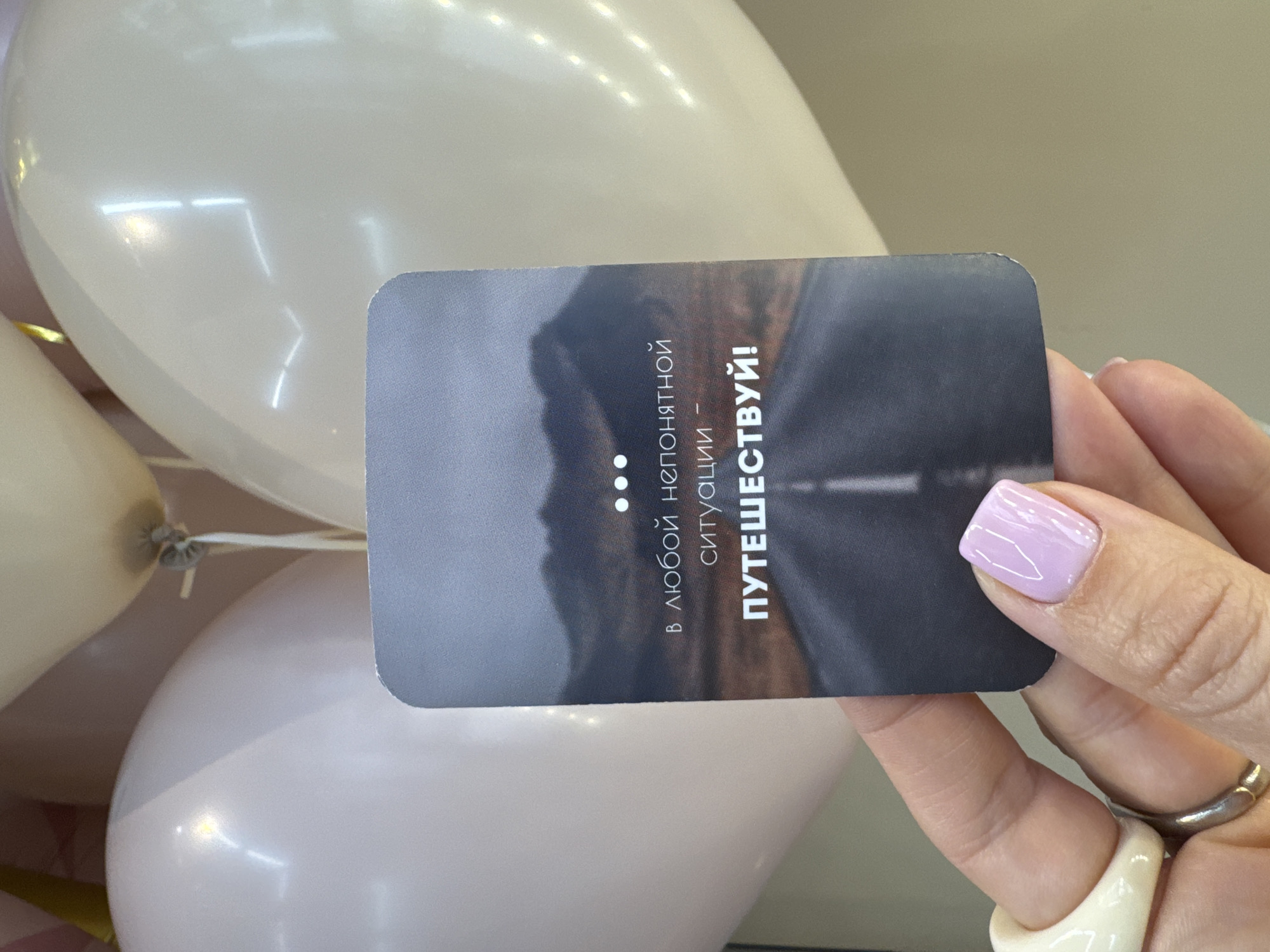 A woman's hand holds a rectangular card against a backdrop of balloons, displaying a road scene transitioning from mountains to asphalt. The card contains Russian text: "В ЛЮБОЙ НЕПОНЯТНОЙ СИТУАЦИИ - ПУТЕШЕСТВУЙ!", which translates to "IN ANY UNCERTAIN SITUATION - TRAVEL!". The balloons appear to be light-colored, possibly white and pink. The woman's hand has a light purple nail polish on one visible fingernail, and she is wearing two rings. The scene's location within Sochi, Russia, cannot be determined from the visual information provided.