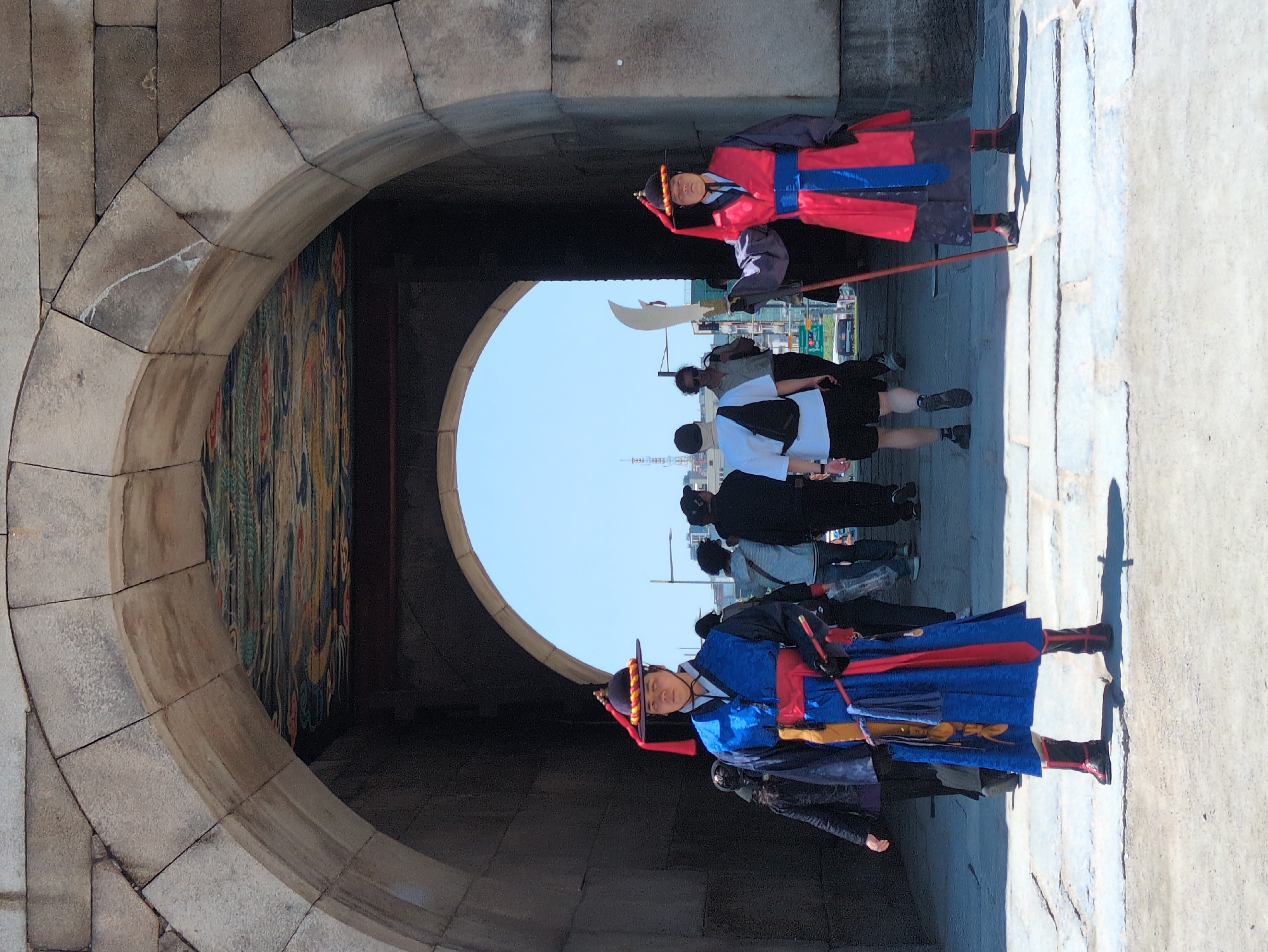 Here is a description of the image:
The image shows a view through a stone archway in Seoul, South Korea. Two men in traditional Korean guard uniforms, one in red and blue, stand on either side of the archway's opening. In the background, people are walking through the open space beyond the arch. The sky is visible as a bright white circle in the distance. A mural decorates one side of the archway. The structure is made of light-colored stone blocks.