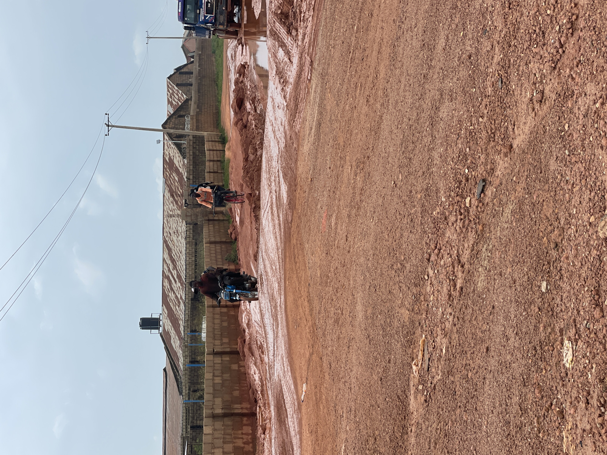 This media file depicts a roadside scene in Jos, Nigeria. The primary subjects are two motorcycles, each carrying at least two people, navigating a muddy, red dirt road. The motorcycles are in motion, suggesting an ongoing journey or commute. The scene is characterized by the rough texture of the unpaved road, which appears to be wet and mucky, likely due to recent rain.
In the background, there are several structures with corrugated metal roofs and brick walls, indicative of residential or commercial buildings. Power lines and utility poles stretch across the sky, suggesting the presence of electricity infrastructure. The sky is mostly clear with a few wispy clouds, hinting at a bright, possibly warm, day. The presence of a large black water tank on a platform further suggests the context of a residential or community area.
The individuals on the motorcycles are dressed in casual clothing. Their expressions are not clearly discernible, but their activity of riding on a challenging road could imply a sense of determination or routine. The overall impression is one of everyday life in a developing urban or semi-urban environment where infrastructure may be less developed. No visible text is present in the media file.