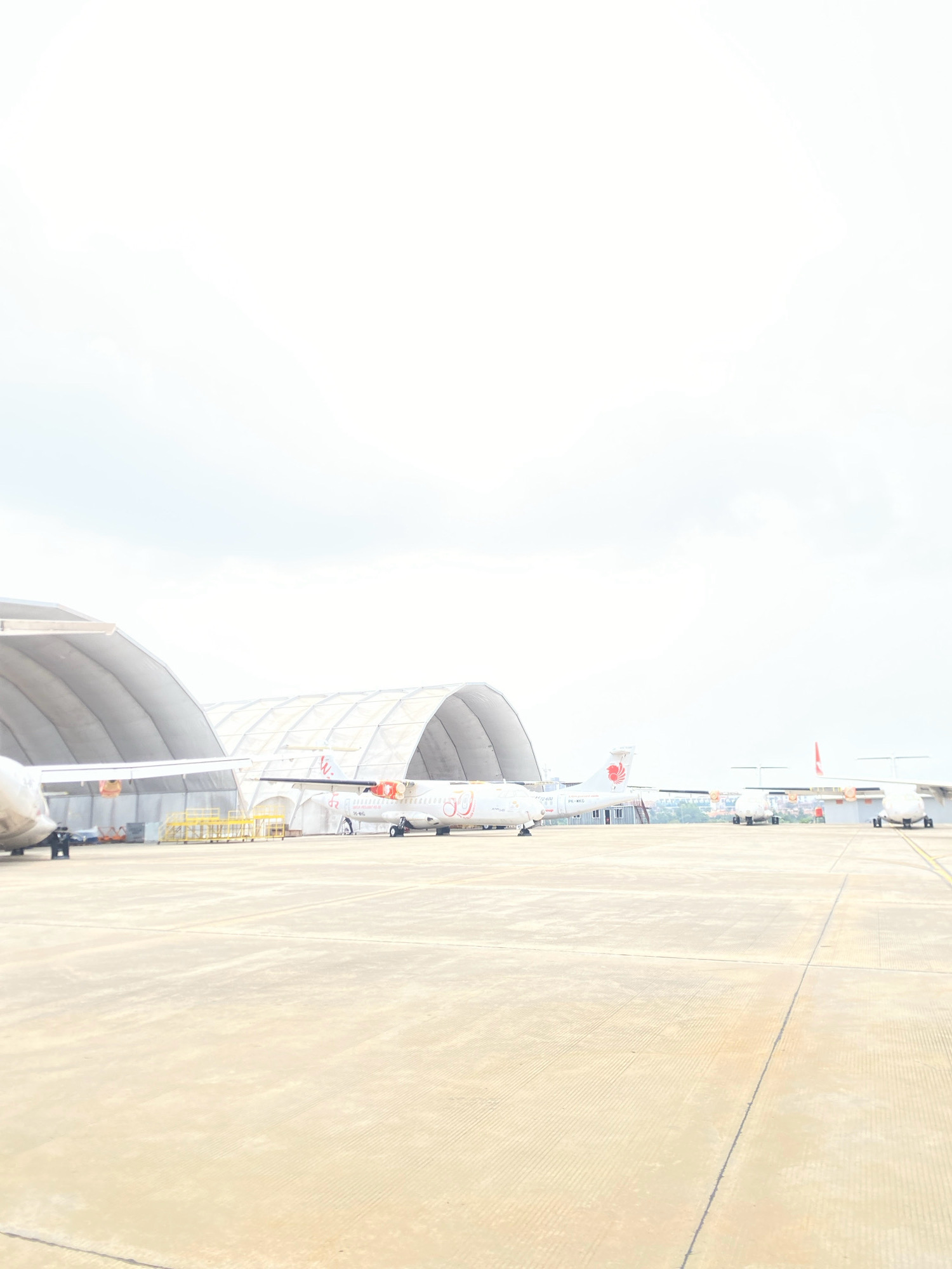 The image shows an aerial view of an airport tarmac in Batam, Indonesia. Several airplanes are parked on the tarmac, some of which are under large, white hangars. The hangars appear to be temporary structures, possibly for aircraft maintenance or storage. The tarmac is made of concrete and shows signs of wear and tear, with visible lines and cracks. In the background, a few buildings and some vegetation can be seen, indicating the presence of a city or town. The sky is overcast, with bright white clouds. The overall scene suggests a busy aviation hub, with aircraft being serviced and prepared for flight.