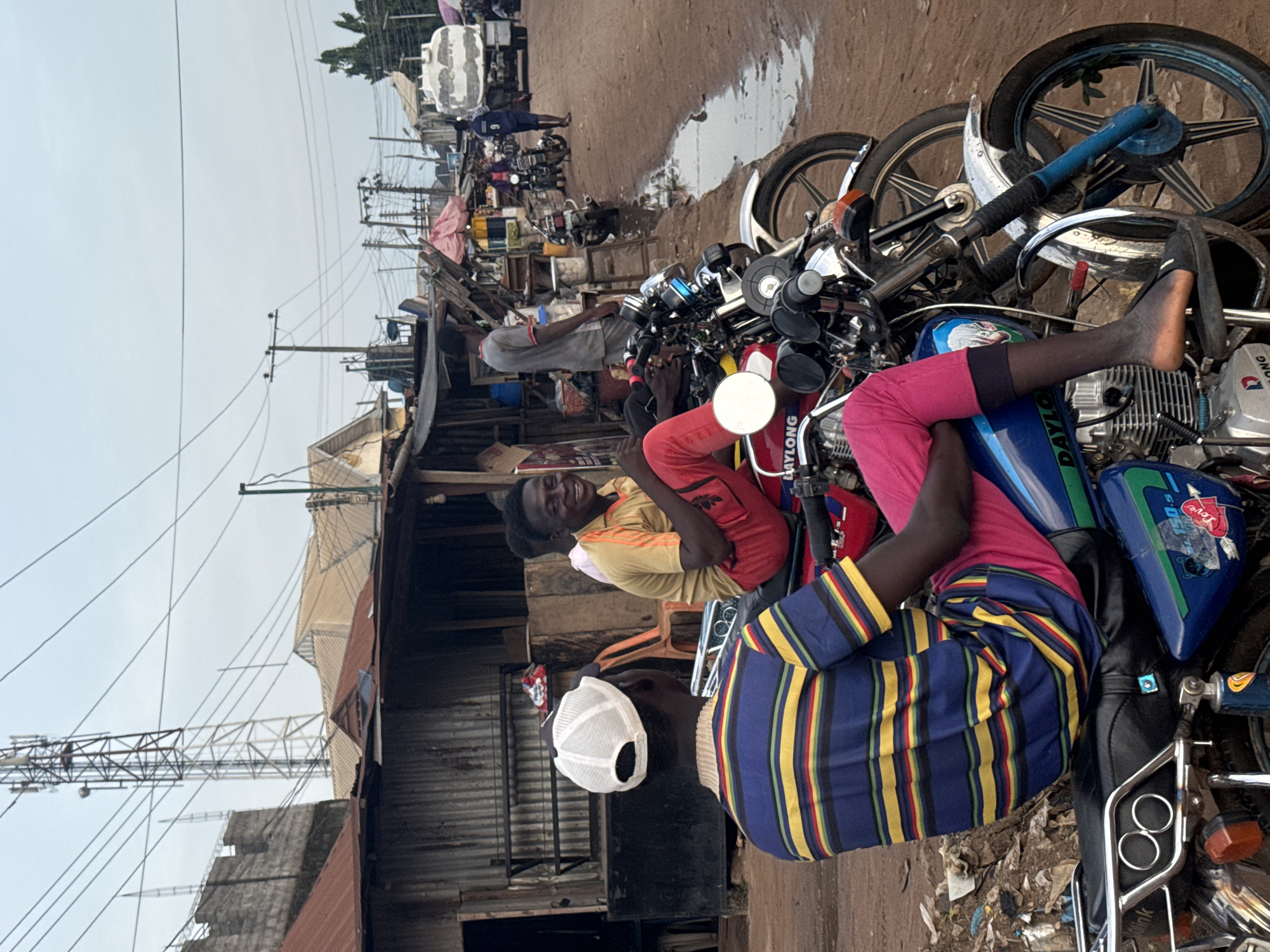 This image captures a lively street scene in Ihiagwa, Nigeria, during the daytime under a clear sky. The photo is oriented vertically but depicts a horizontal street.
In the foreground, several individuals are gathered around motorcycles on a dirt road. A man with his back to the viewer wears a blue and yellow striped shirt and a white baseball cap, sitting on a motorcycle. Beside him, another man with a broad smile looks towards the camera, sitting on a red motorcycle and wearing a yellow t-shirt and red pants. The legs of a third person, dressed in pink pants, are visible, resting casually on the tank of a blue and green motorcycle. The brand "DAYLONG" is clearly visible on the fuel tanks of several motorcycles, and a small heart sticker with "Love" is seen on the blue motorcycle. The ground is unpaved, appearing dusty and mixed with mud or debris, with some puddles of water visible.
The midground features a bustling street lined with market stalls or small shops constructed from corrugated iron sheets and wooden frames. Other people are visible moving or standing among the structures. Utility poles with numerous power lines crisscross the sky above the buildings. In the background, further down the street, taller metallic structures, possibly communication towers, are visible against the bright sky. The scene depicts a typical busy day in a street market or thoroughfare.