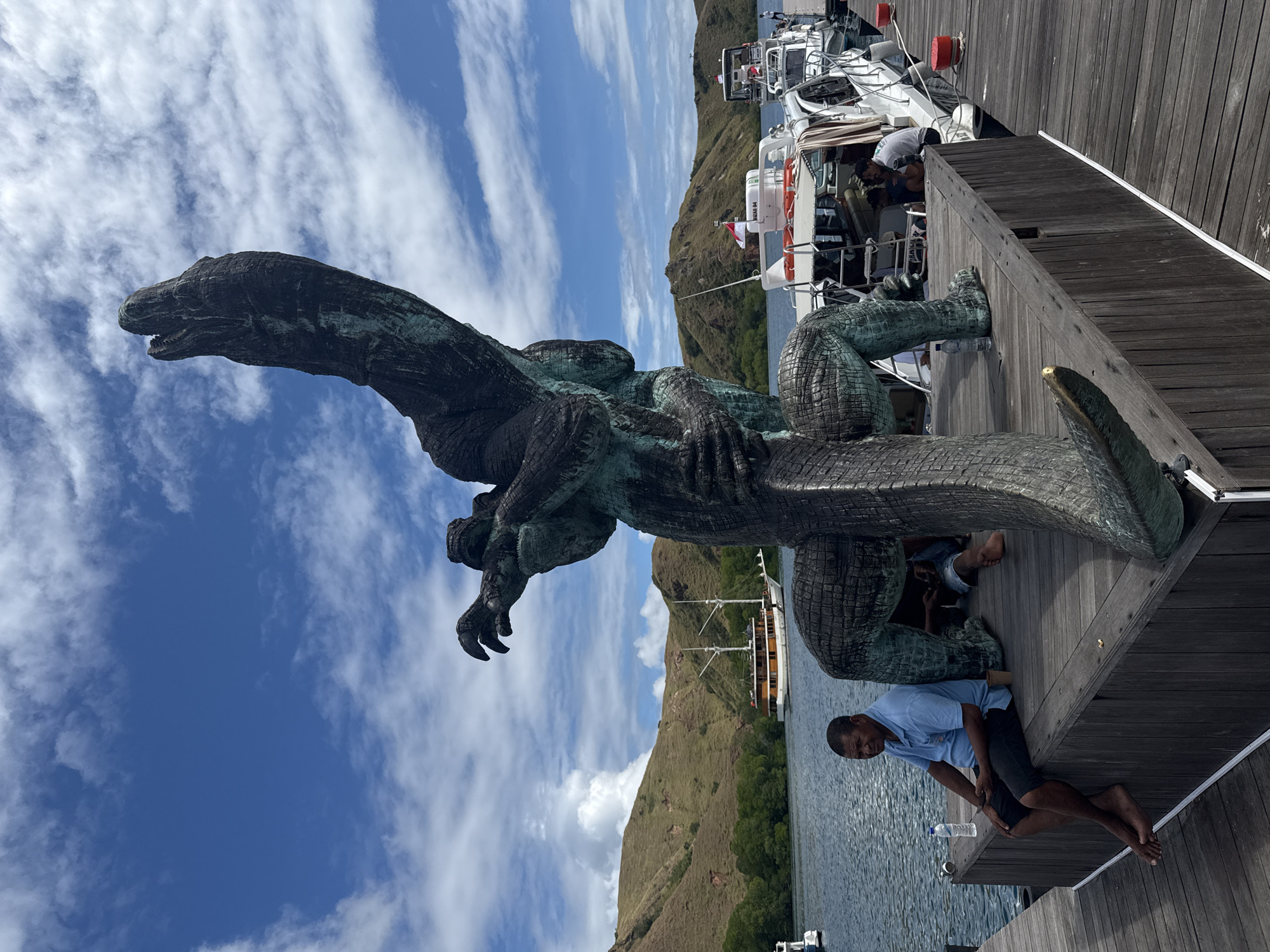 Here is a detailed description of the image:
A horizontally oriented medium shot depicts a bronze crocodile statue erected on a wooden pier. The statue stands out in intricate detail against a vibrant backdrop of a partly cloudy sky, a tranquil sea, and an undulating mountainous terrain.
The crocodile statue dominates the foreground, its scales and form sculpted with precision. A person sits on the wooden pier, positioned next to the statue. The individual appears to be posing or resting. Behind the crocodile statue and to the right of the frame, several boats are moored at the pier.
The backdrop reveals an expansive body of water reflecting the sky above. On the opposite shore, a landscape of rolling mountains covered with vegetation creates a distinct horizon line. The sky is bright blue, adorned with streaks of white clouds that suggest a pleasant, sunny day.
The photograph seems to have been taken in Komodo Island, Indonesia. The statue is located on a pier. The picture appears to be taken during daylight under bright sunlight.