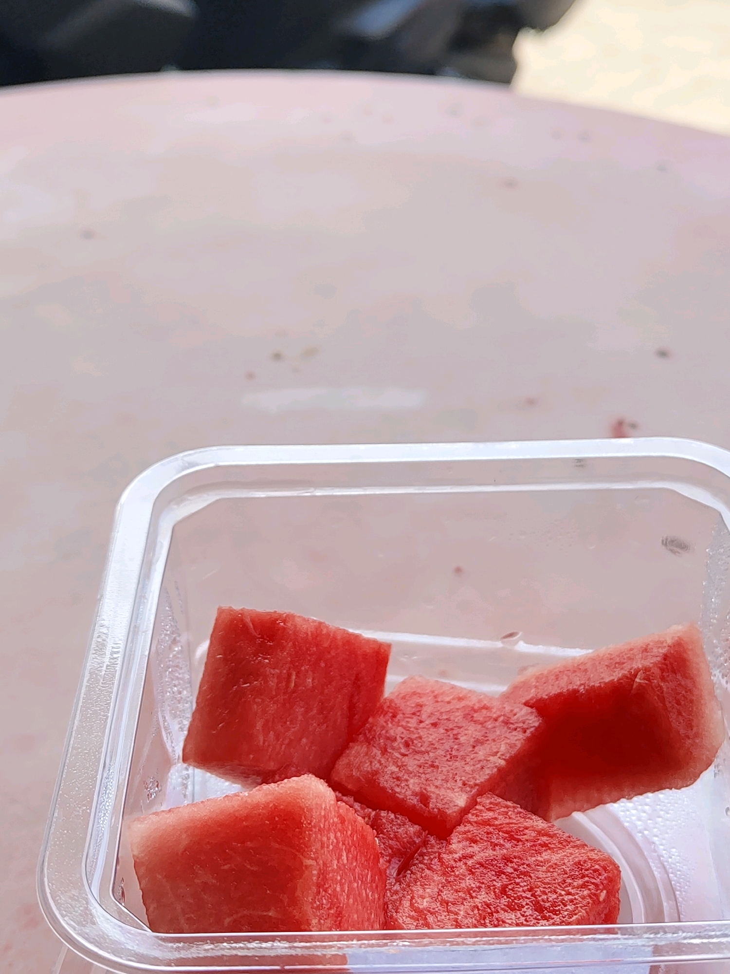 A close-up, low-angle shot shows several cubed pieces of ripe watermelon inside a clear plastic container. The watermelon pieces are a vibrant red and appear juicy. The container has a slightly frosted texture on its interior, suggesting condensation. The background is out of focus, but appears to be a light-colored, textured surface, possibly a table. The top of the image shows a blurred dark object, perhaps a person's clothing or an item in the background. The scene appears to be outdoors, as indicated by the soft, natural lighting. There is no discernible location within the city of Probolinggo, Indonesia, that can be identified from this image.