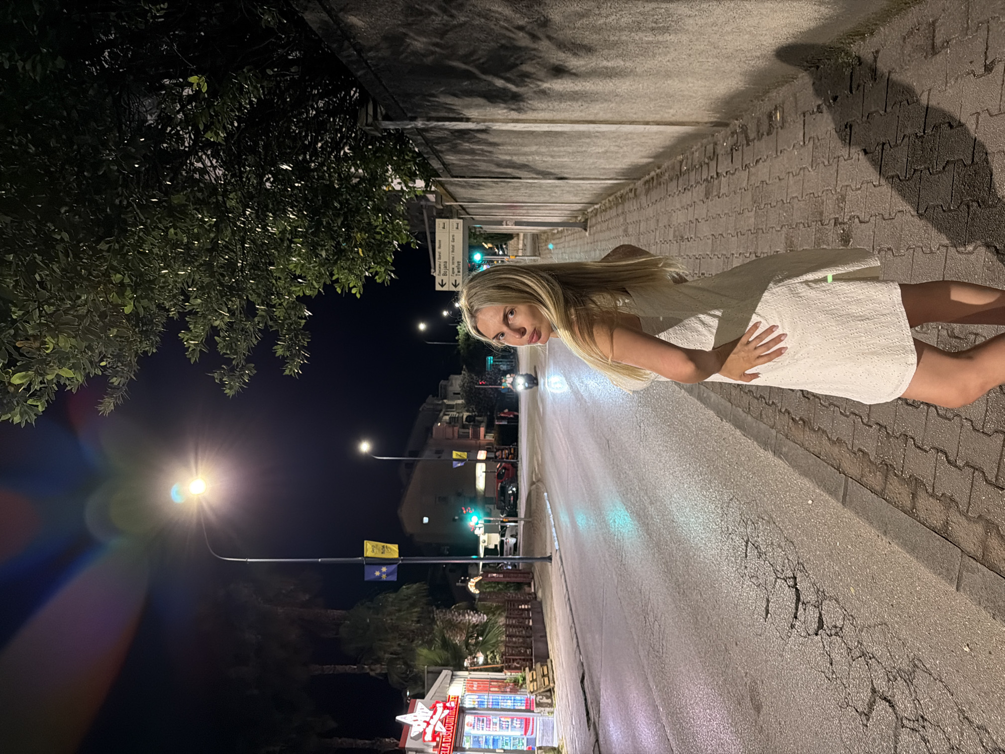 A woman with blonde hair, wearing a white dress, is leaning towards the camera on a sidewalk next to a street at night. She is looking at the camera with a slight smile. The street has traffic lights, a few vehicles, and streetlights that illuminate the scene. The sidewalk is made of interlocking pavers. In the background, there are buildings and trees. A signpost with directional arrows and text is visible on the sidewalk. There is a store with a bright sign in the distance. The time of day is night, indicated by the darkness and artificial lighting. The weather appears to be clear. The scene is in Budva, Montenegro, and the woman's pose and expression suggest a casual and relaxed mood.