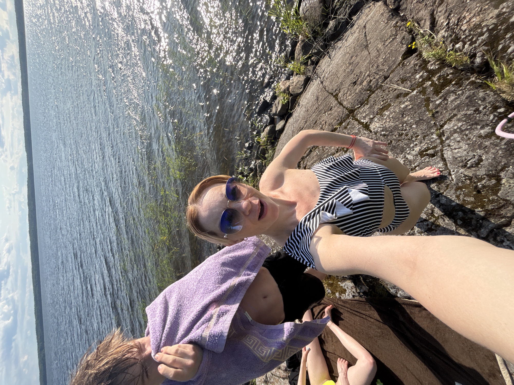 In Shuiskaya Chupa, Russia, at a lakeside location, a group of people are gathered on the rocky shore. A young person, partially obscured by a purple towel, stands near the water's edge. A woman wearing a black and white striped swimsuit and sunglasses is in the foreground, holding a camera and appearing to take a selfie. Other individuals' legs are visible, indicating they are also present on the shore. The lake's surface is rippled, reflecting sunlight, and vegetation grows along the waterline. The scene depicts a casual outdoor gathering.