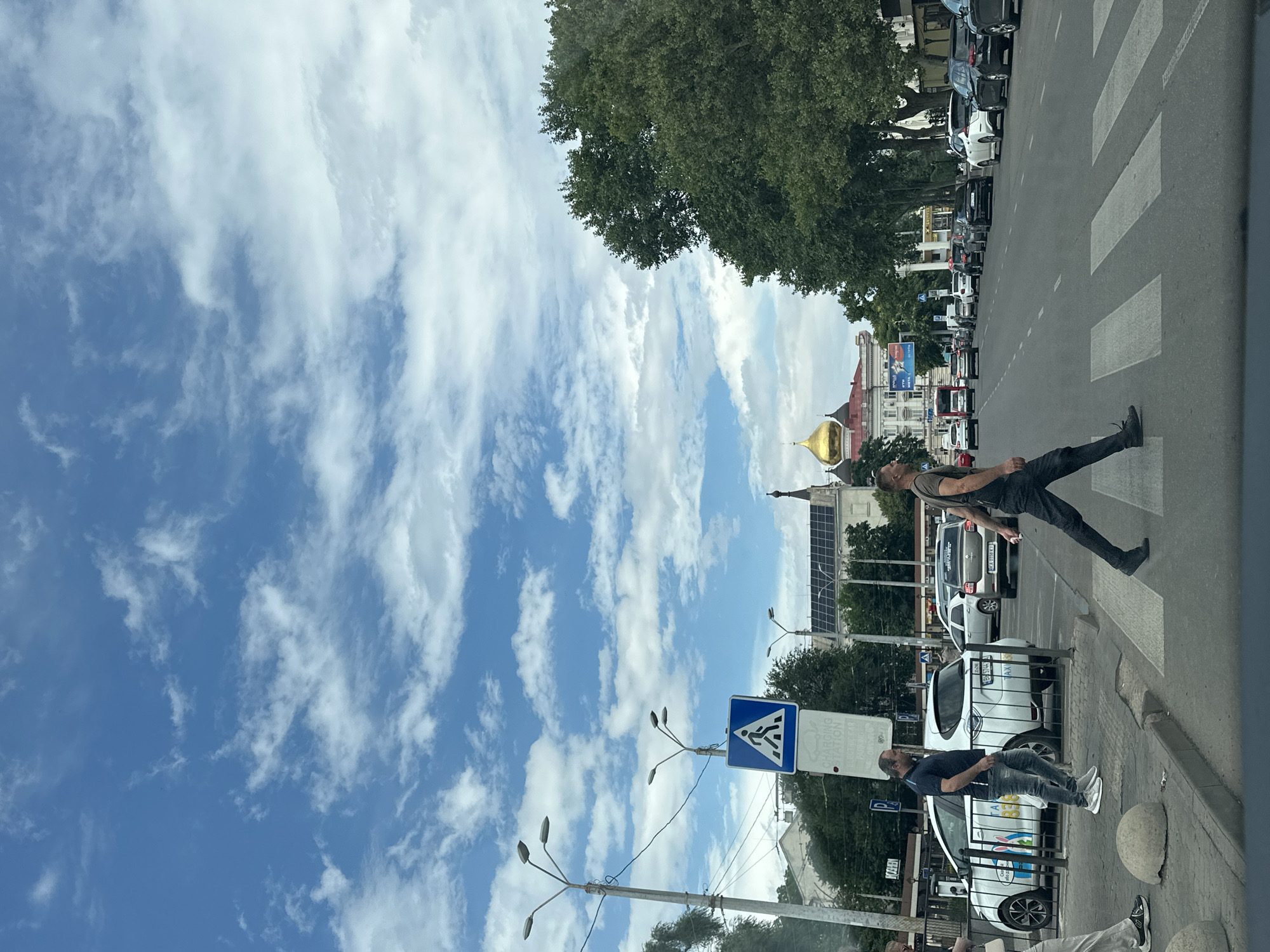 The image captures a view of the intersection of Deribasivska Street and Preobrazhenska Street in Odesa, Ukraine, on a cloudy day. A pedestrian crossing is visible in the foreground, with several people walking across it. A man in a dark t-shirt and jeans is walking from left to right, while another man in a dark t-shirt and black pants is walking from right to left.
Several cars are also present on the street, including a white electric taxi with "TAXI" written on its side and a silver car with a Ukrainian license plate. In the background, a large building with a golden dome, likely the Transfiguration Cathedral, stands out. Trees line the street, and a blue sky with white clouds is visible above. A street lamp and a pedestrian crossing sign are also present on the left side of the image. The overall scene depicts a typical urban street in Odesa.