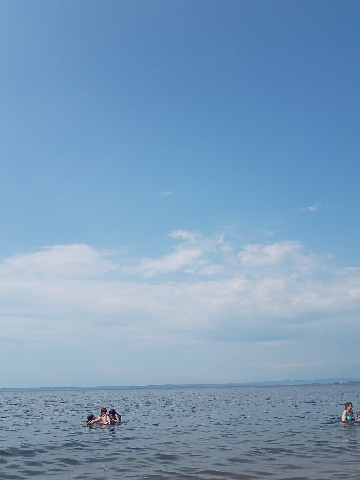 A wide shot captures a serene body of water under a bright blue sky with scattered clouds. The water is calm with gentle ripples, reflecting the sky. In the middle distance, a group of three individuals are gathered on a float, appearing to be interacting or relaxing. Further to the right, a lone individual in a swimsuit is visible in the water. In the far distance, a hazy horizon line suggests land or a distant coastline. The scene evokes a sense of leisure and a warm, pleasant day at the beach.