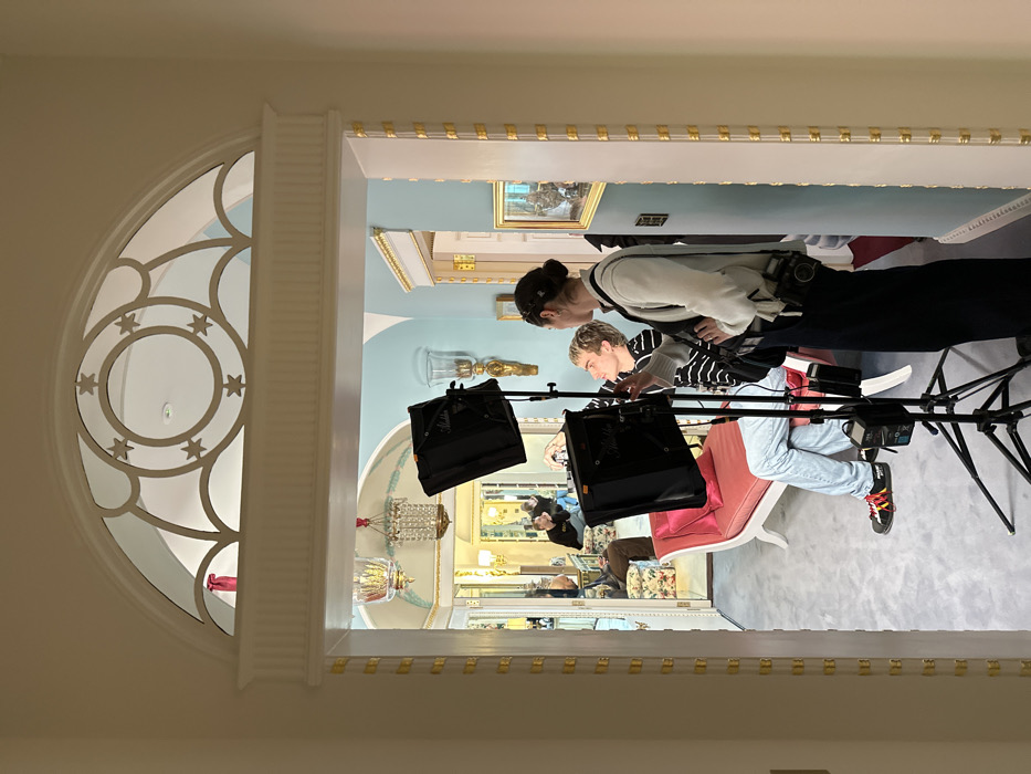 The image depicts an indoor scene within the opulent interior of Annabel's, a private members' club located in Berkeley Square, Mayfair, London, United Kingdom. The view is framed by a grand white arched doorway, distinguished by a semi-circular transom featuring a star-and-circle motif and a fluted architrave with gold accents.
Through the doorway, a professional photoshoot or video shoot is in progress on a light gray carpeted floor. In the immediate foreground, a female, observed from a three-quarter back angle, stands to the right, wearing a white cardigan, dark trousers, and a black cross-body bag with a medium-format camera attached to her left hip. She is facing left, seemingly directing or interacting with a younger individual with short, light-colored hair, seated on a white chaise lounge upholstered in pink velvet. This individual wears a black and white striped long-sleeve top and light blue jeans, with black sneakers featuring red and yellow laces, and gestures with their right hand.
Positioned between these two individuals is a black photography light stand supporting two "Aladdin" brand LED light panels, illuminating the seated subject.
Beyond the immediate setup, the lavish interior of Annabel's extends into multiple connected rooms. These rooms feature light blue walls adorned with intricate gold moldings and trim, complemented by elaborate white architectural details. Hanging crystal chandeliers and decorative wall sconces with glass shades contribute to the luxurious atmosphere. Further into the background, reflections in multiple mirrors reveal additional indistinct figures and richly decorated spaces, including another person seated on a patterned armchair and one figure wearing a yellow top visible in a distant mirror.