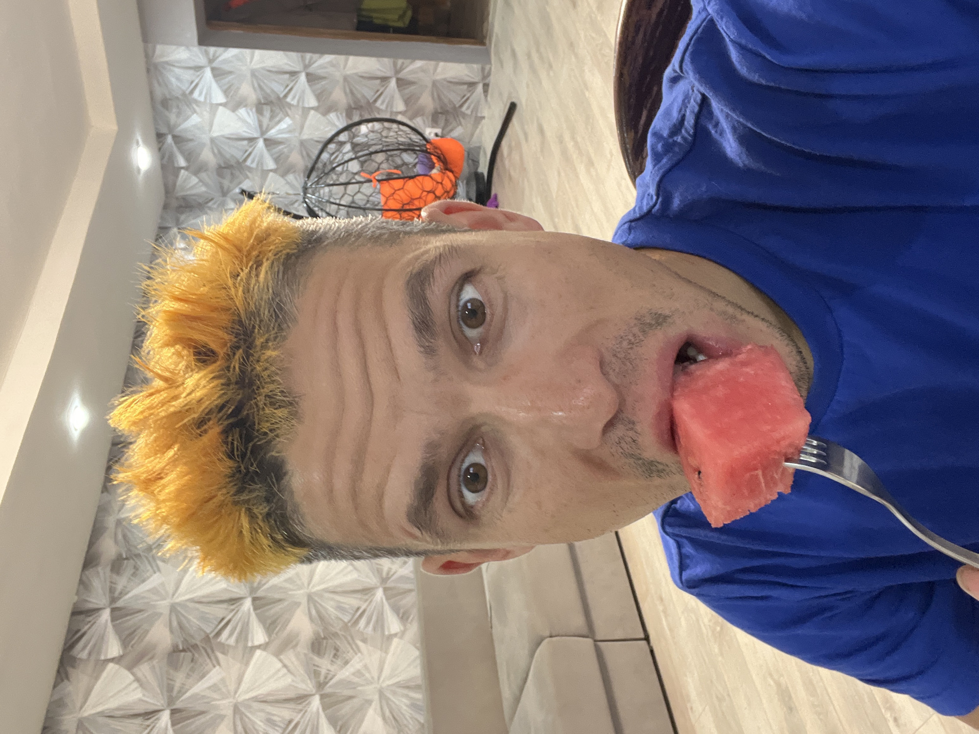 A close-up, vertically oriented selfie shows a person with brightly dyed orange hair eating a slice of watermelon from a fork. The person is wearing a blue t-shirt. Their eyes are open and looking at the camera, and a slice of watermelon is held to their mouth on a silver fork. The background features a patterned wall and a wooden floor, suggesting an indoor setting. A glimpse of a black cage-like object with orange accents is visible behind the person's head. The lighting appears to be artificial, indicating it could be daytime or nighttime. The overall expression suggests a casual, perhaps humorous, moment. The location context is Chaikovsky, Russia.
