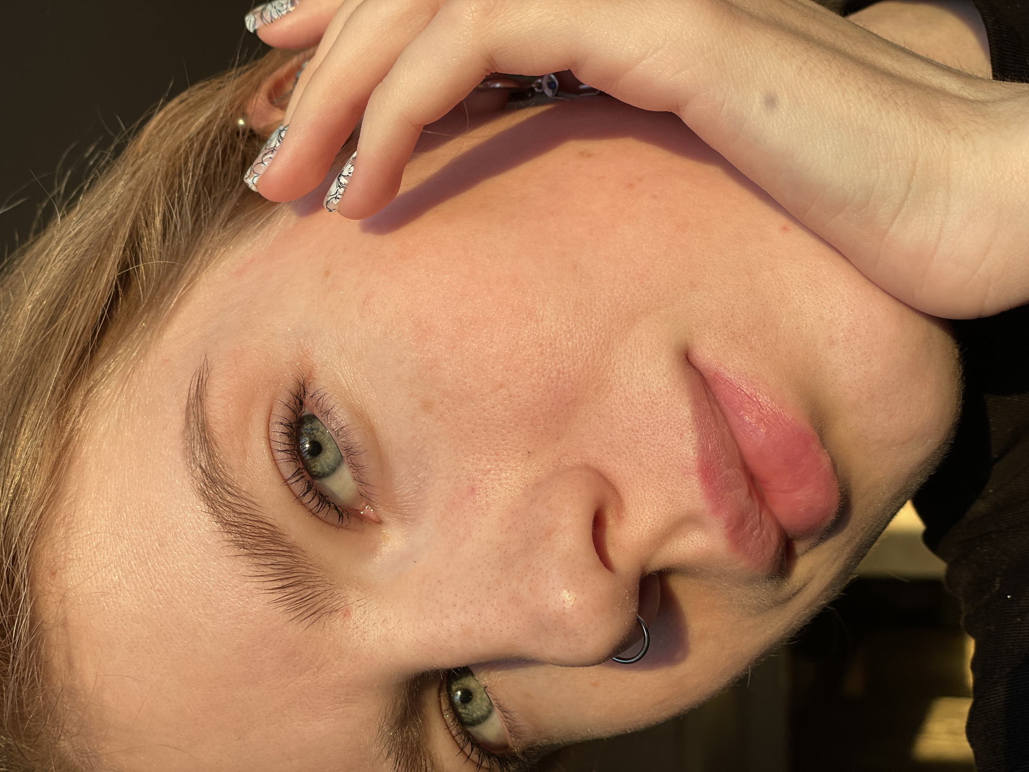 A close-up, high-angle portrait captures the face of a person with fair skin, light blonde or light brown hair, and striking blue-green eyes. The subject's head is tilted and rotated in the frame, with their face illuminated by strong, warm, directional light, characteristic of golden hour (morning or late afternoon/early evening). Their left hand, adorned with intricate white and black patterned fingernails, rests gently on their head/hair, partially obscuring their forehead.
The person's skin displays natural texture with subtle freckles. Prominent eyelashes and natural, somewhat bushy eyebrows frame their clear, observant eyes. A silver septum piercing is visible on their nose, and their lips are a natural pinkish hue, slightly parted. A small amount of body hair is visible on the hand/forearm resting near the face. The overall expression is calm and neutral, conveying a sense of relaxation or contemplation.
The background is indistinct and dark, suggesting an indoor setting or a deeply shadowed area. The image provides no visible text or specific location cues, though the context indicates the location is Izhevsk, Russia. The composition highlights the subject's facial features and the interplay of light and shadow, creating an intimate and artful portrayal.