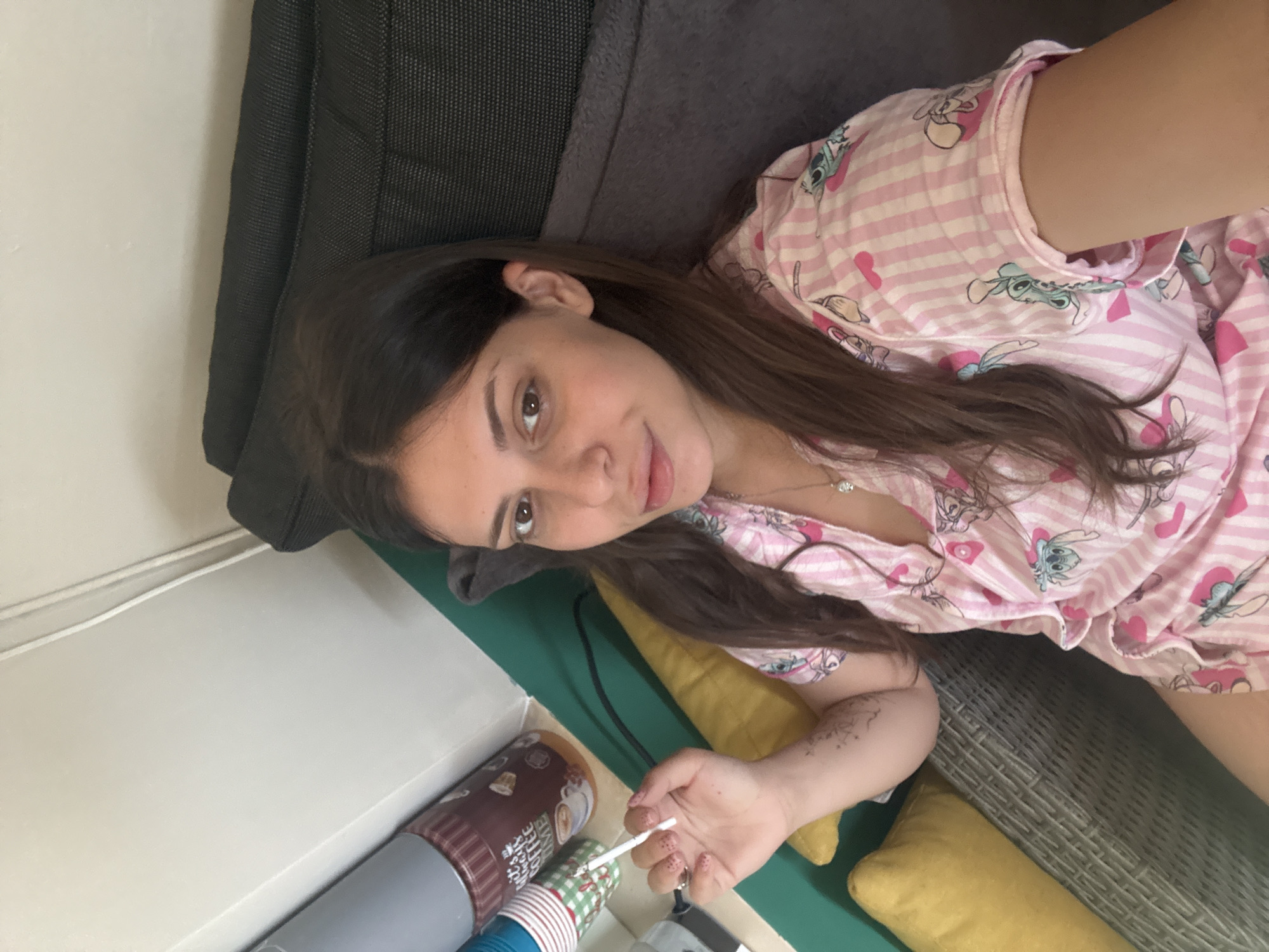 A woman is holding a cigarette in her right hand, her left arm resting on a yellow pillow. She is wearing pink and white striped pajamas with cartoon characters. She is seated on a dark gray cushion with a woven chair visible beneath. In the background, to the left, are two cylindrical containers and a stack of paper cups. The scene appears to be indoors or on a covered patio, given the lighting and wall texture. The overall impression is casual and relaxed. No specific location in Plovdiv, Bulgaria can be determined from the image.