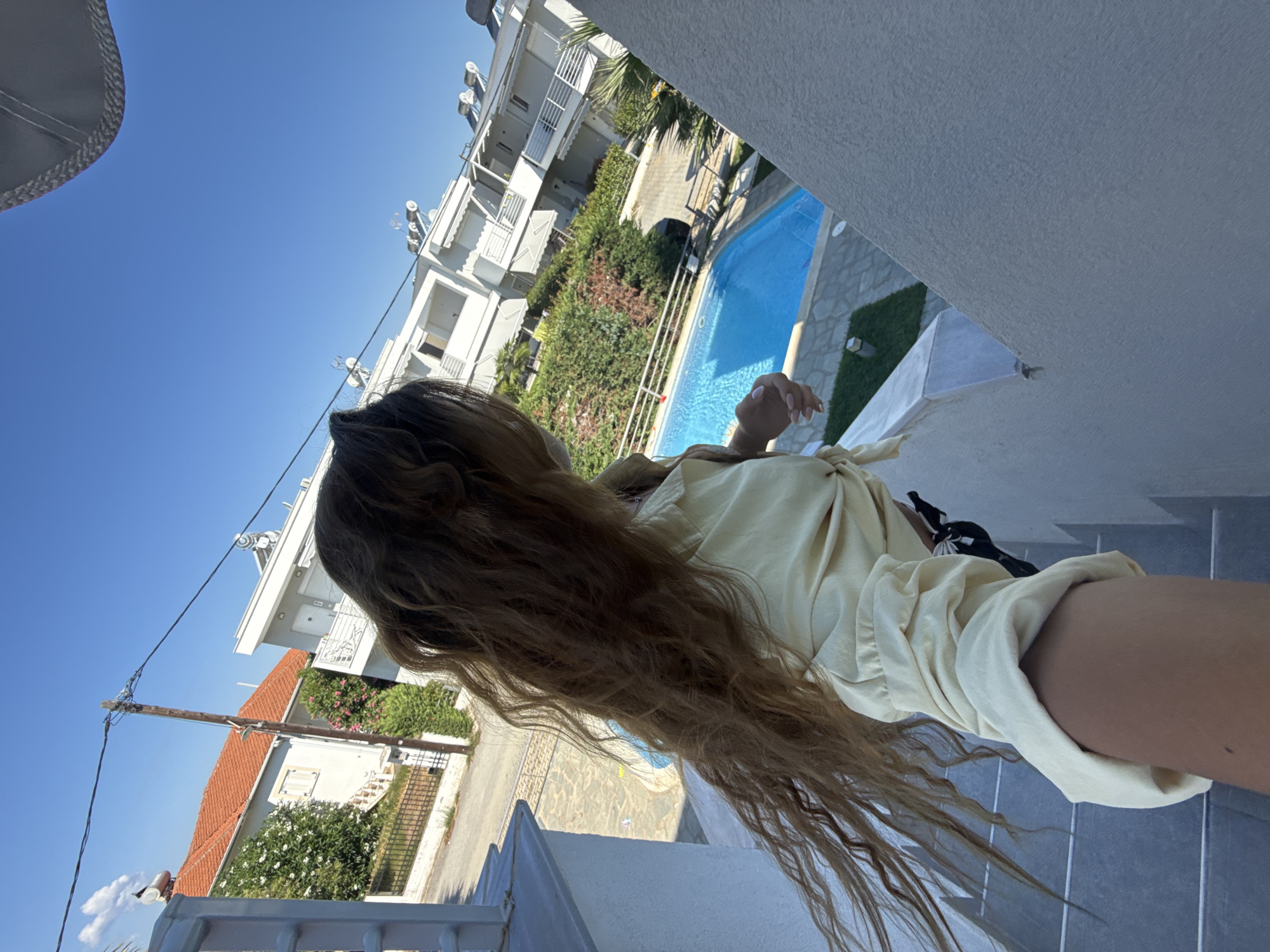 A young woman with long, brown hair is standing on a balcony overlooking a swimming pool and houses in Ofrynio Beach, Greece. She is wearing a light yellow blouse and black shorts. The woman is holding a smartphone, taking a selfie. The houses are modern, with white facades and balconies. The swimming pool is blue and surrounded by a paved patio. The sky is clear and blue. The image is taken from a high angle, looking down at the scene.