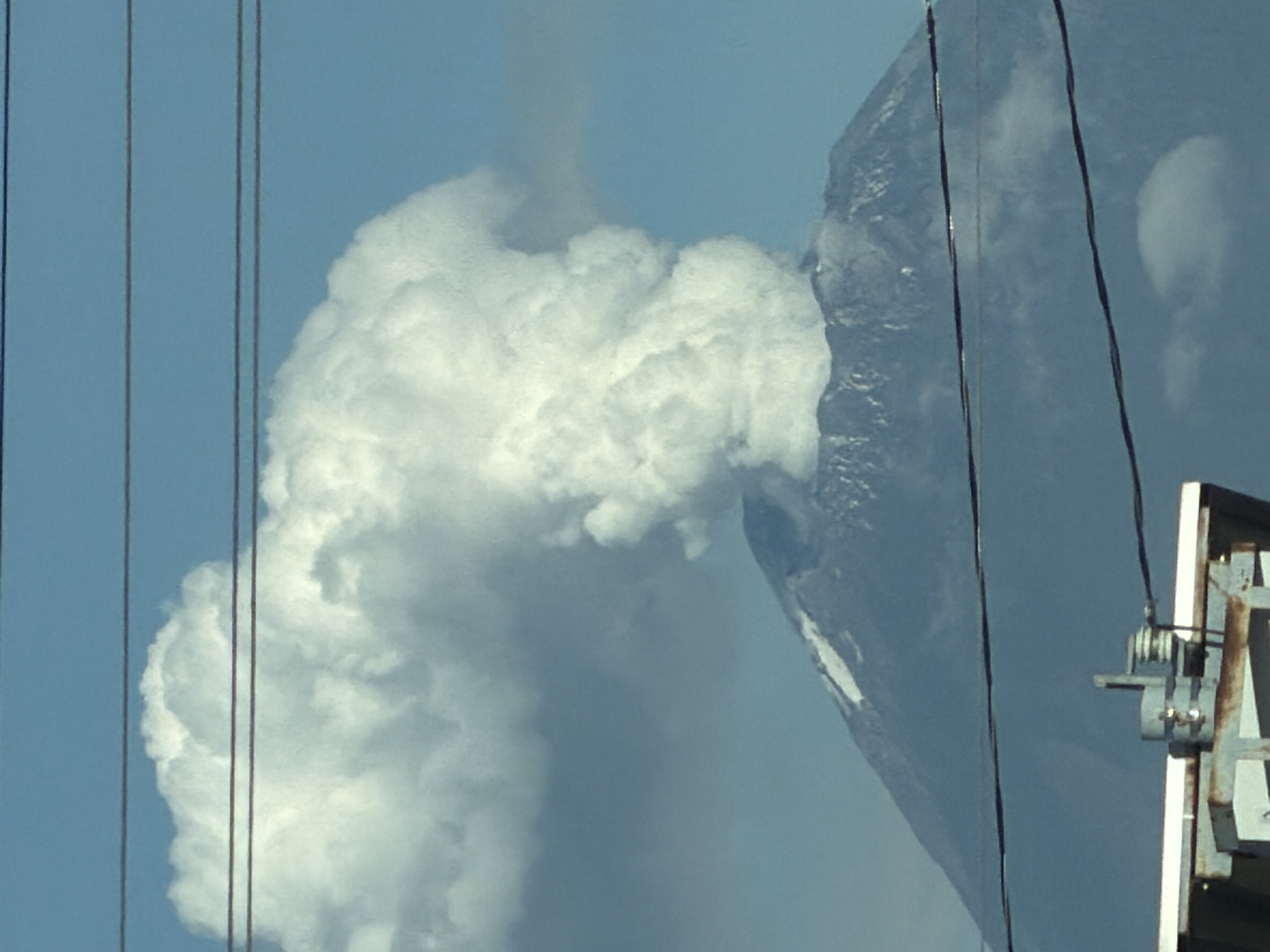 The image shows a wide shot of a volcano erupting, with a large plume of smoke and ash billowing into the sky. The volcano is located in San Bernardino Tlaxcalancingo, Mexico, a municipality in the state of Puebla. The foreground of the image is occupied by power lines and a portion of a structure that appears to be part of a utility pole or some form of electrical equipment. The volcano itself dominates the background, with its slopes visible beneath the massive eruption cloud. The sky is a clear blue, providing a stark contrast to the dark smoke. The scene suggests an active and potentially dangerous natural event.