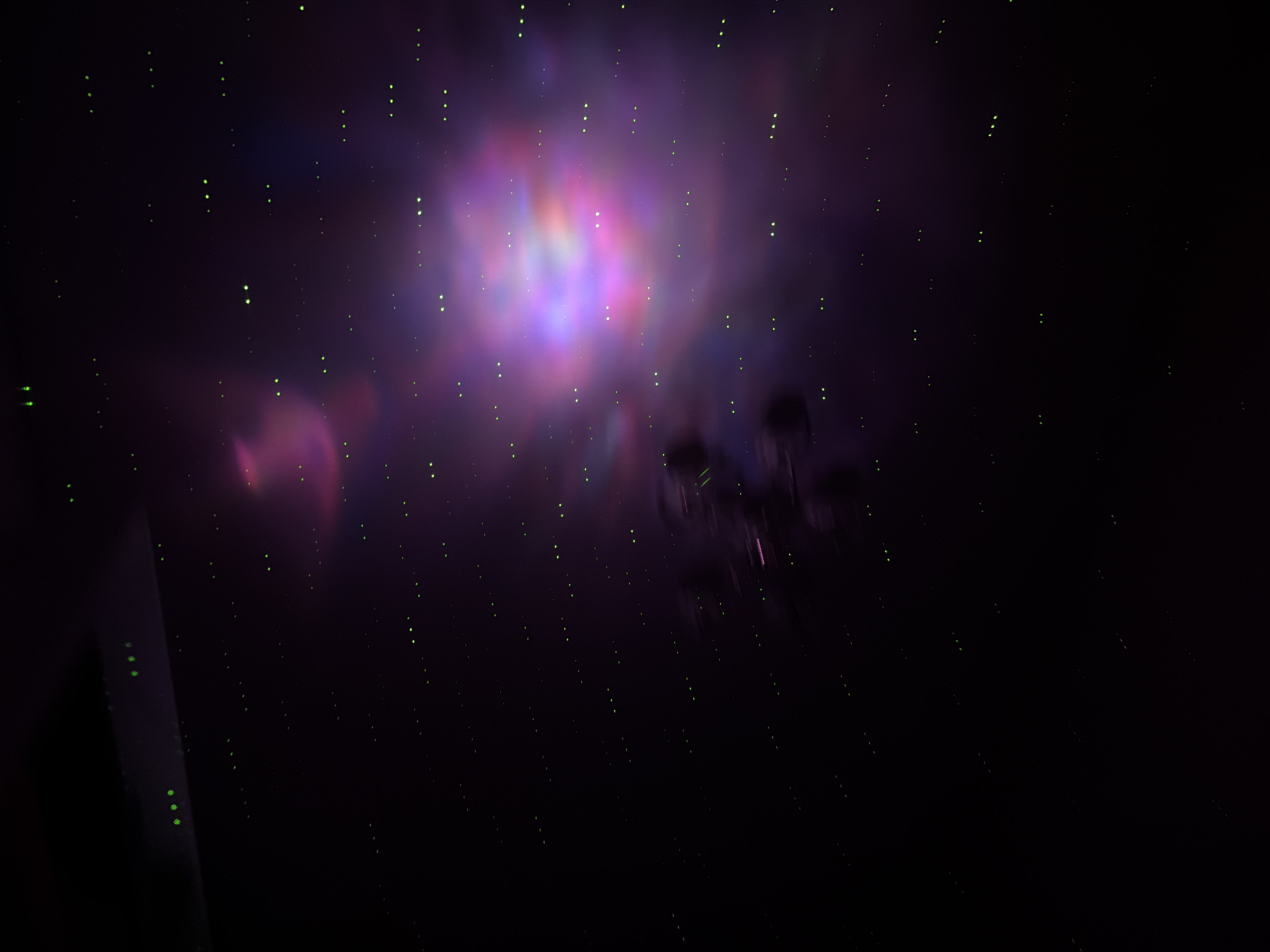 The image depicts a dark environment with a bright, colorful nebula-like formation in the upper center. The colors are predominantly purple, blue, and pink. Scattered throughout the darkness are numerous small, green dots, resembling stars. In the lower left corner, a dark, angular structure with a few green dots is visible. In the mid-right, some indistinct dark shapes appear, possibly resembling objects or figures, but are too blurry to identify. The image appears to be an artistic representation or a simulated view of a nebula with elements of an urban setting due to the linear structure and the presence of regular green dots, which might be artificial lights. I lack the information to provide the specific location in the city of Perm, Russia.