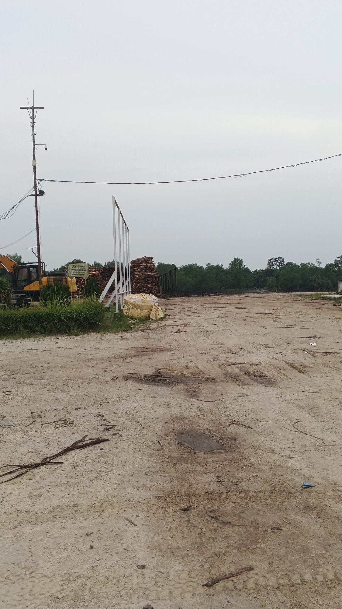 This is a vertical, eye-level, outdoor, wide shot of a logging yard on an overcast day. The foreground is a dirt lot with tire tracks, scattered debris, and a small puddle. In the midground, to the left, a yellow excavator is parked. A tall, metal pole with electrical wires, a security camera, and an antenna stands next to it. To the right of the excavator is a stack of lumber, a white metal gate structure, and two large, yellow tarp-wrapped bundles. In the background, a dense line of green trees forms the horizon. The sky is a uniform, light gray. This scene is located in Merempan Hilir, Indonesia.