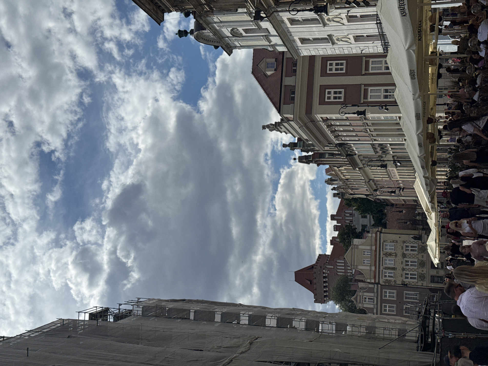 This image depicts a view of Stary Rynek, the Old Market Square in Poznan, Poland, on a cloudy day. The foreground shows a crowd of people mingling and seated at outdoor cafes under white umbrellas, some of which bear the text "KSIĄŻĘCE". To the left, a tall building is partially covered in white sheeting and scaffolding, indicating ongoing construction or renovation. Further into the scene, a red-roofed, turreted building, likely a historic tower, is visible amidst a cluster of architectural styles of the surrounding market square buildings. The sky is a mix of blue patches and substantial grey clouds.